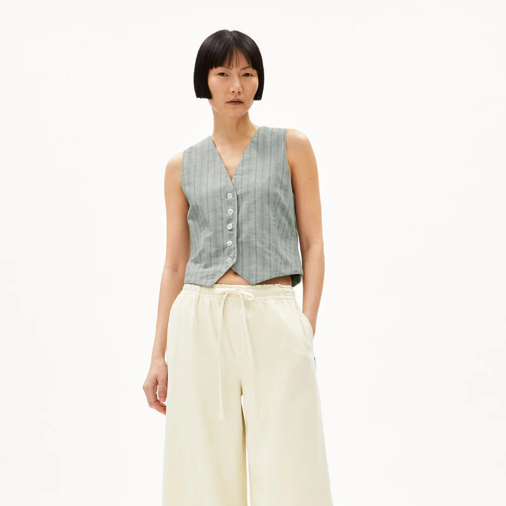 Eine Person mit kurzen schwarzen Haaren trägt eine ärmellose, hellgraue, geknöpfte Weste mit Nadelstreifen und eine weite, cremefarbene Hose mit hoher Taille, dazu schwarze Slide-Sandalen, die vor einem einfarbig weißen Hintergrund stehen.