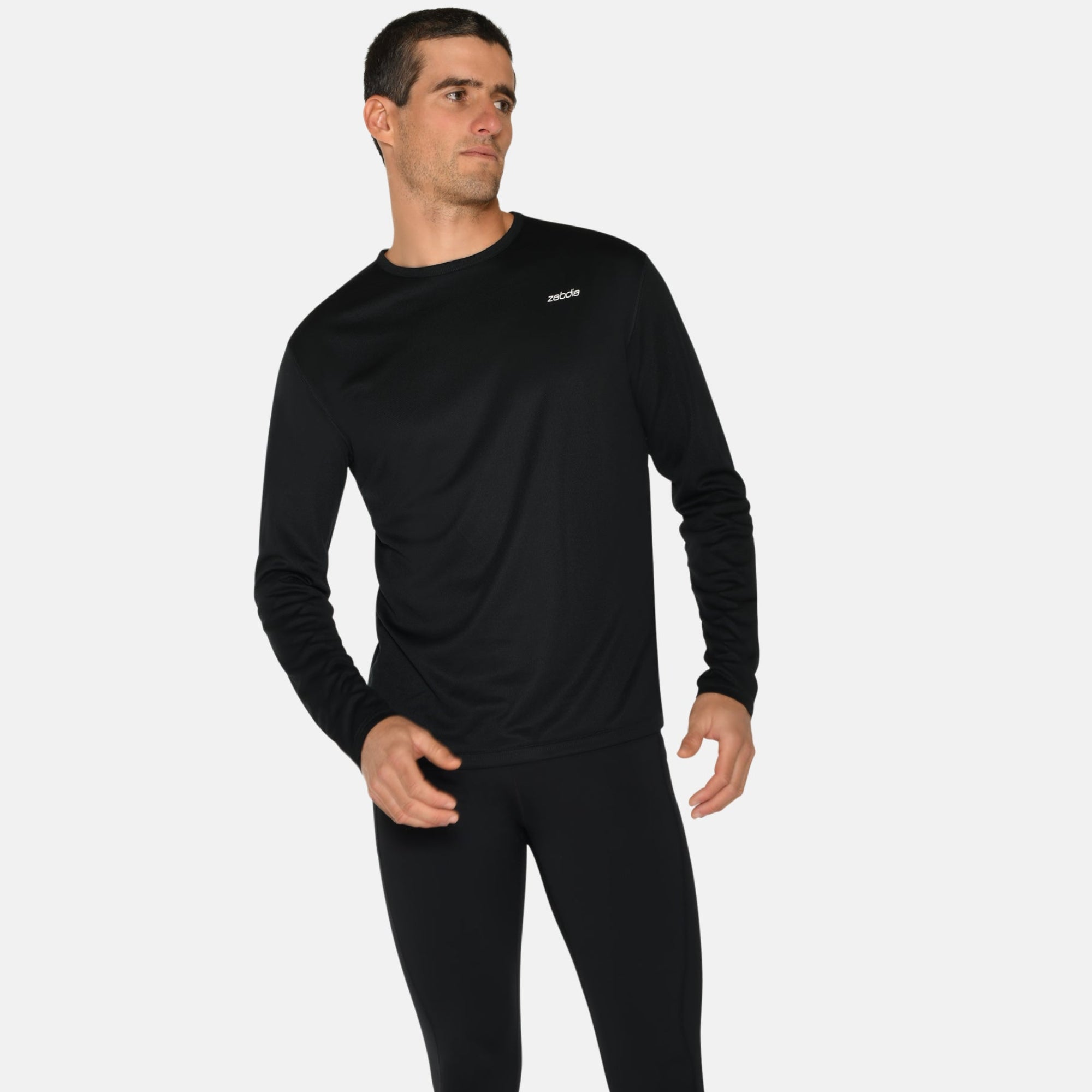 Ein Mann steht vor einem weißen Hintergrund und trägt ein schwarzes langärmeliges Sporthemd, schwarze Leggings und blaue Laufschuhe. Er blickt leicht zur Seite und hält eine Hand in der Nähe seiner Taille und die andere an seinem Oberschenkel.