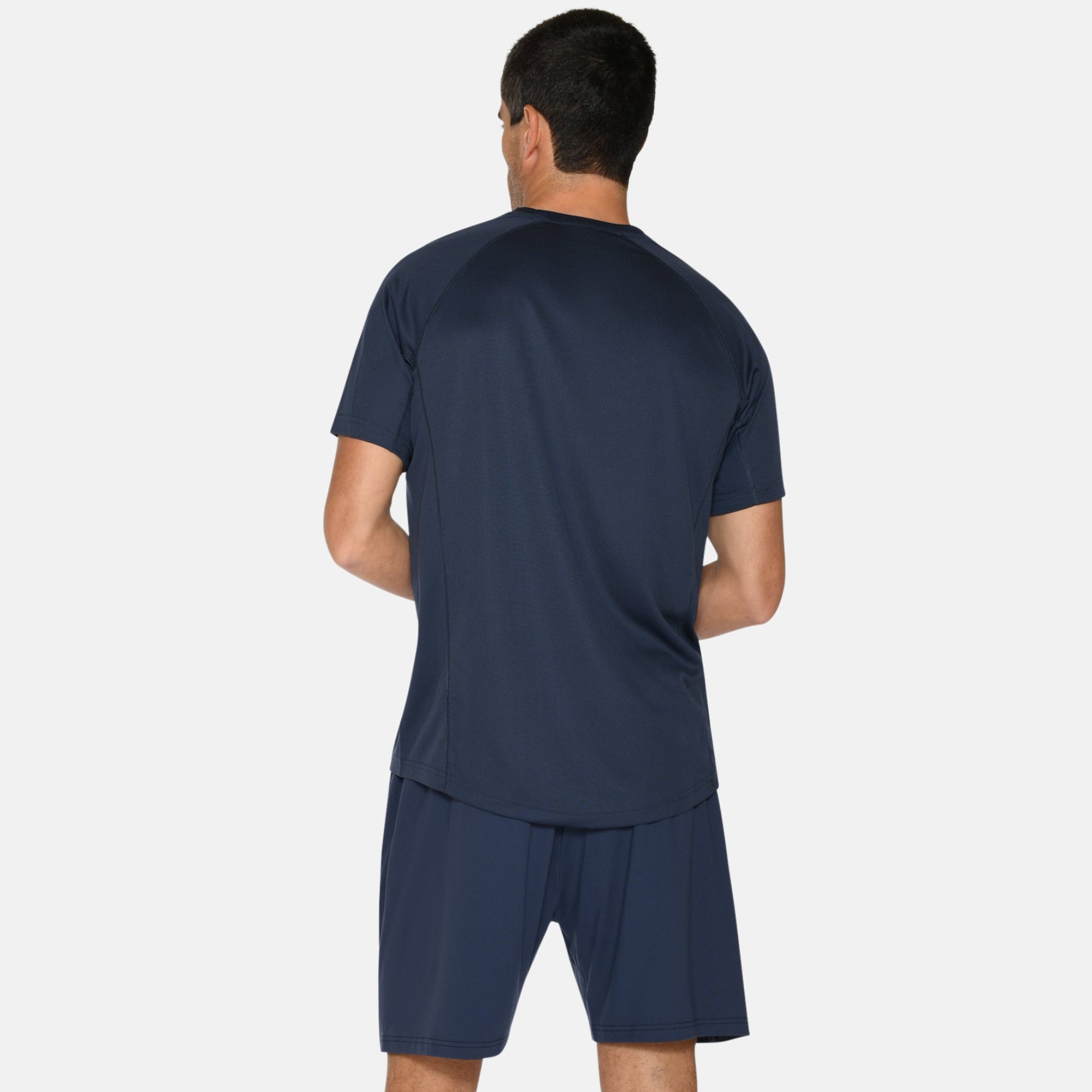 Ein Mann steht mit dem Gesicht zur Kamera, trägt ein marineblaues Sporthemd und Shorts, gestreifte marinefarbene Socken und blaue Laufschuhe mit orangefarbenen Akzenten. Der Hintergrund ist einfarbig weiß.