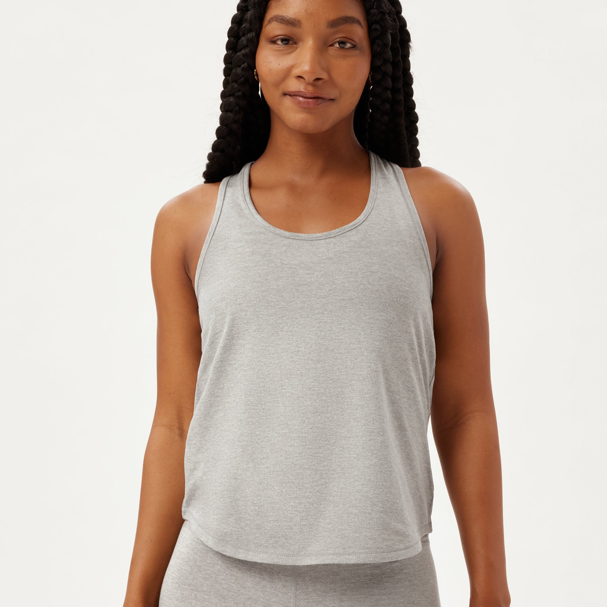 Eine Frau mit langen, geflochtenen Haaren trägt ein ärmelloses, hellgraues Tank-Top und eine passende Hose. Sie steht vor einem schlichten weißen Hintergrund und lächelt sanft in die Kamera.