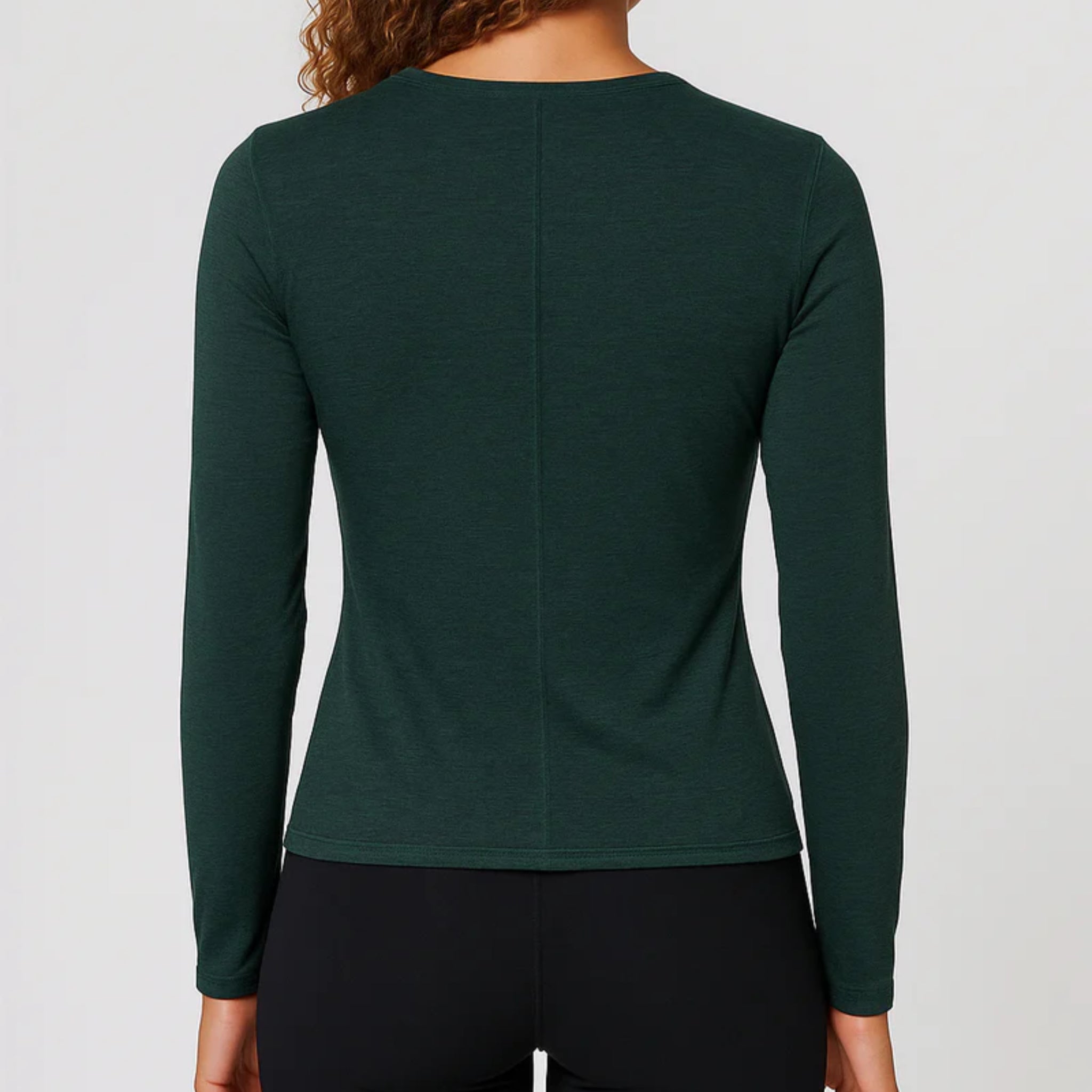 Gezeigt wird eine Person mit lockigem Haar von hinten, die ein langärmeliges, dunkelgrünes Oberteil und eine schwarze Hose vor einem schlichten hellen Hintergrund trägt.