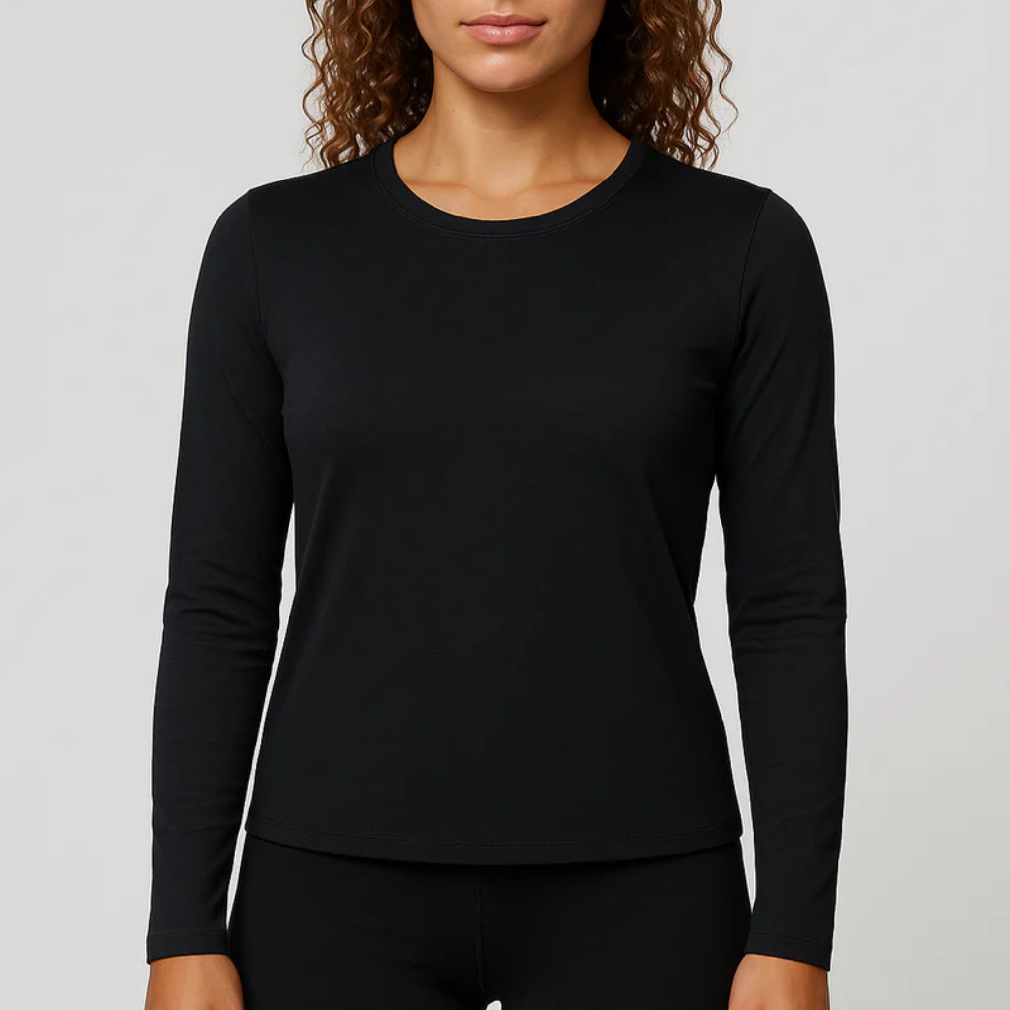 Eine Frau mit lockigem Haar, die ein schlichtes schwarzes langärmeliges Hemd und eine schwarze Hose trägt, steht vor einem neutralen Hintergrund. Ihr Gesicht ist teilweise sichtbar, und ihre Arme liegen entspannt an ihren Seiten.