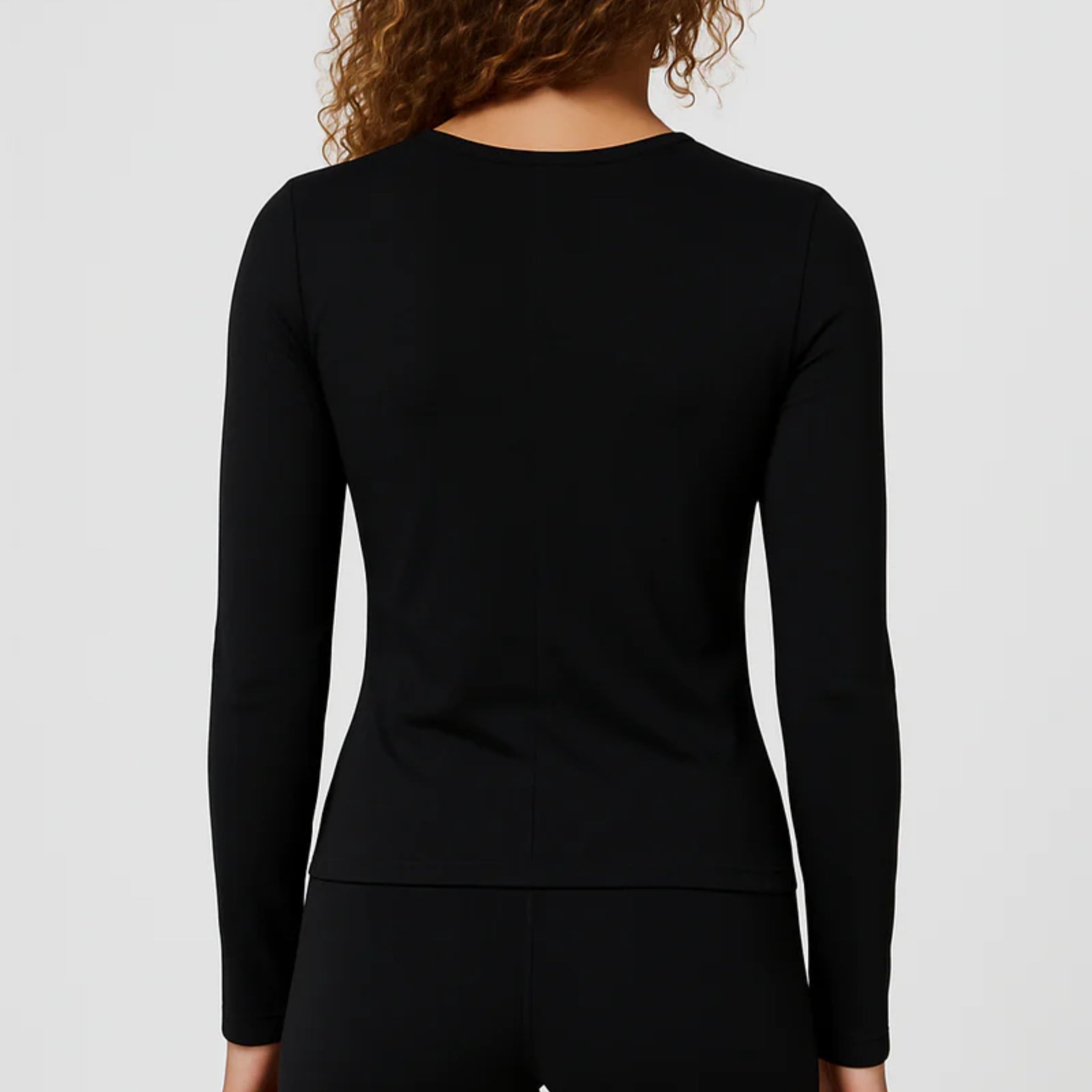 Eine Person mit lockigem Haar, die ein schwarzes, langärmeliges Oberteil und eine dazu passende schwarze Hose trägt, von hinten vor einem schlichten, hellen Hintergrund dargestellt.