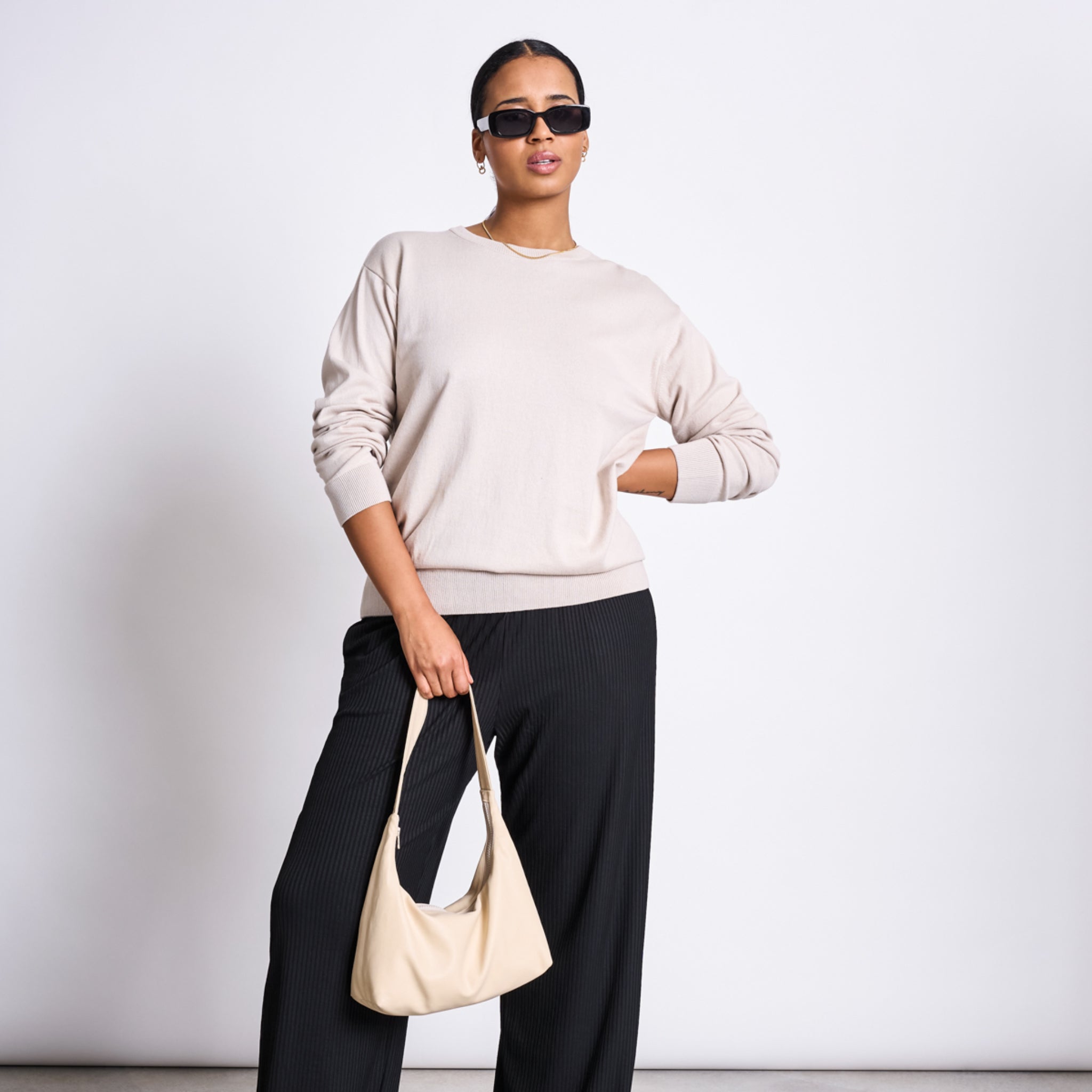 Frau in beigem Pullover und schwarzer Hose trägt eine Sonnenbrille und hält eine hellbeige Handtasche.