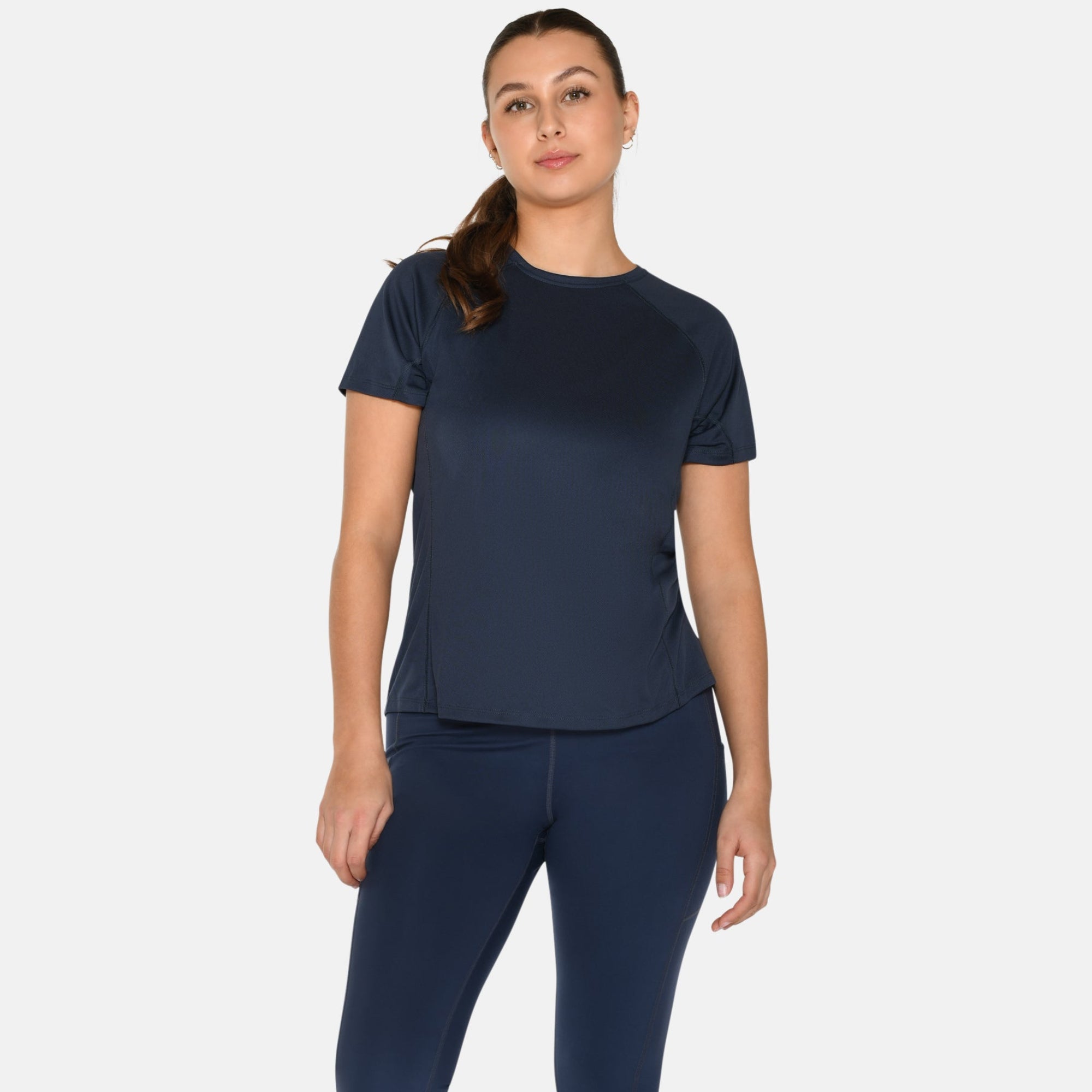 Eine Frau steht vor einem schlichten weißen Hintergrund und trägt ein marineblaues kurzärmeliges Sportoberteil, passende marineblaue Leggings und weiße Turnschuhe. Sie trägt ihr Haar zu einem Pferdeschwanz und hat einen neutralen Gesichtsausdruck.