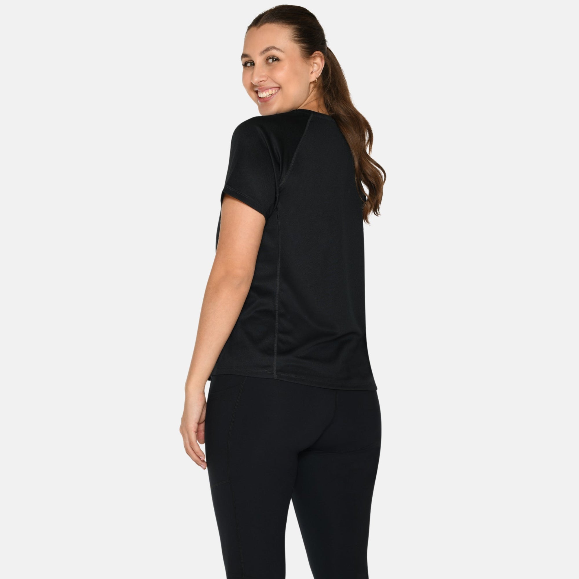 Eine Frau mit langen braunen Haaren in einem Pferdeschwanz, die ein schwarzes kurzärmeliges Oberteil, schwarze Leggings und weiße Turnschuhe trägt, steht vor einem schlichten hellen Hintergrund und lächelt über ihre Schulter.