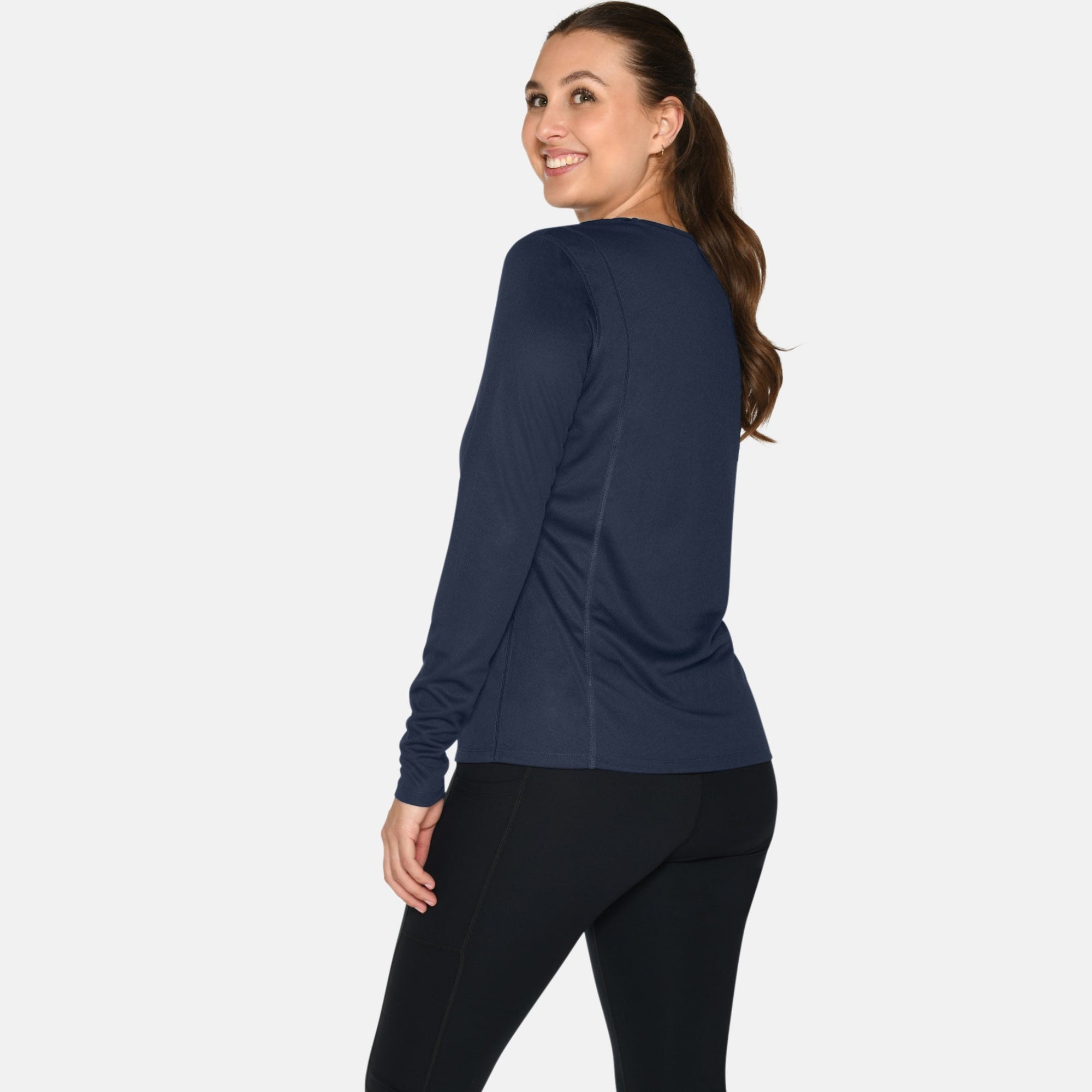 Eine Frau steht mit dem Gesicht zur Seite und schaut lächelnd über ihre Schulter. Sie trägt ein marineblaues langärmeliges Oberteil, schwarze Leggings und weiße Turnschuhe vor einem schlichten weißen Hintergrund.