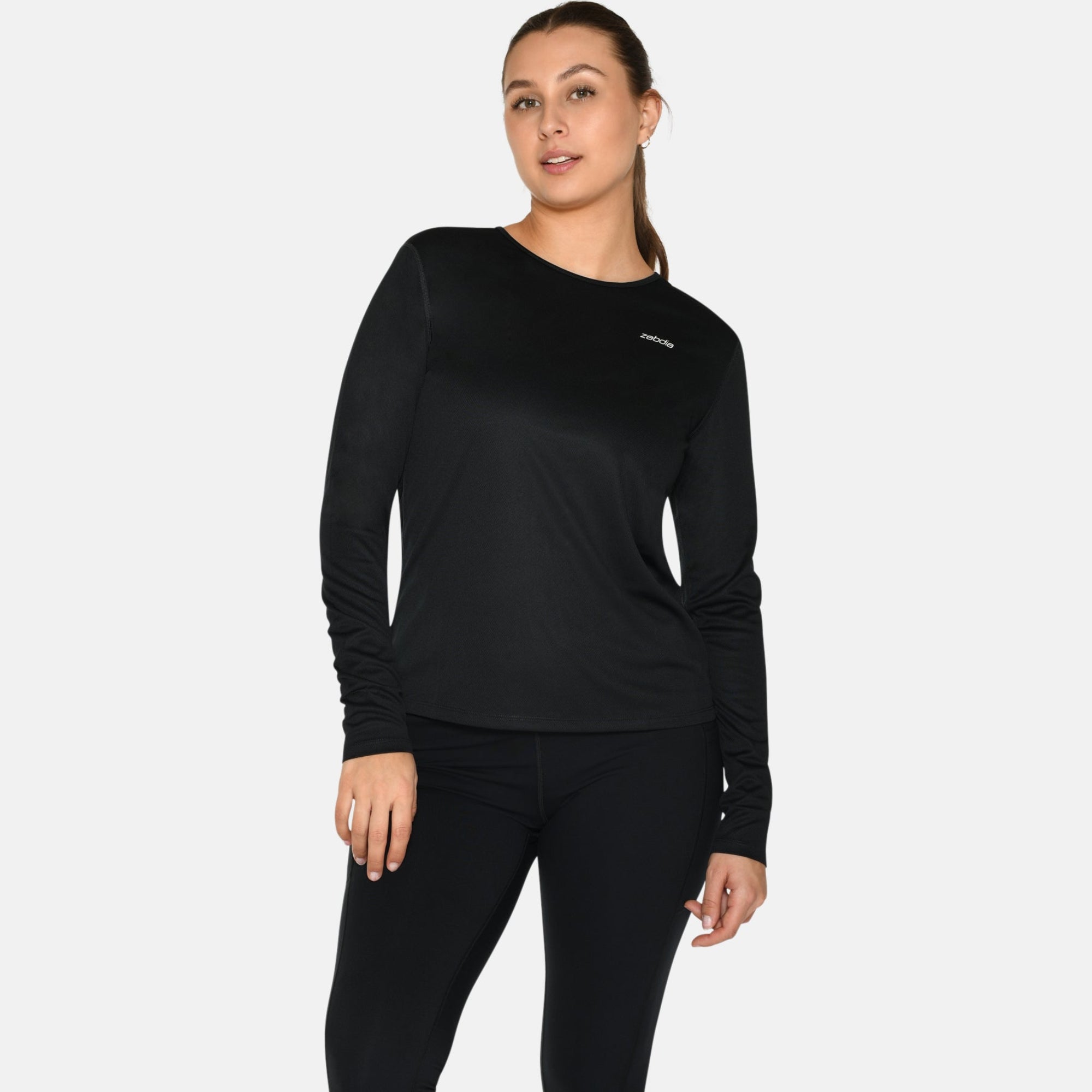 Eine Frau steht mit dem Gesicht nach vorne, trägt ein schwarzes langärmeliges Oberteil, schwarze Leggings und weiße Turnschuhe vor einem schlichten weißen Hintergrund.