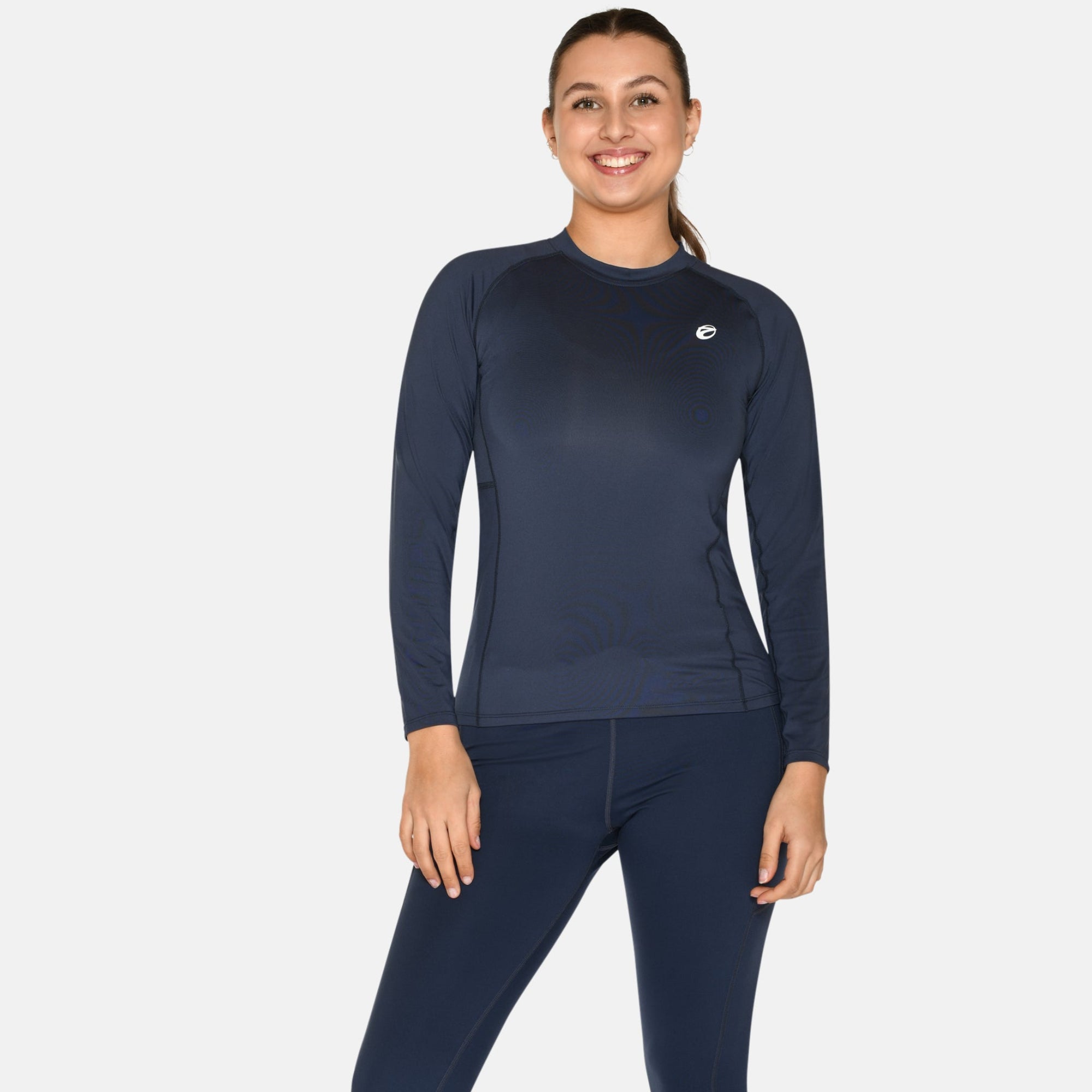 Eine Frau, die ein marineblaues, langärmeliges Sportoberteil und passende Leggings trägt, steht lächelnd mit einer Hand an der Seite und der anderen auf ihrem Bein, vor einem schlichten weißen Hintergrund. Sie trägt außerdem weiße Turnschuhe.