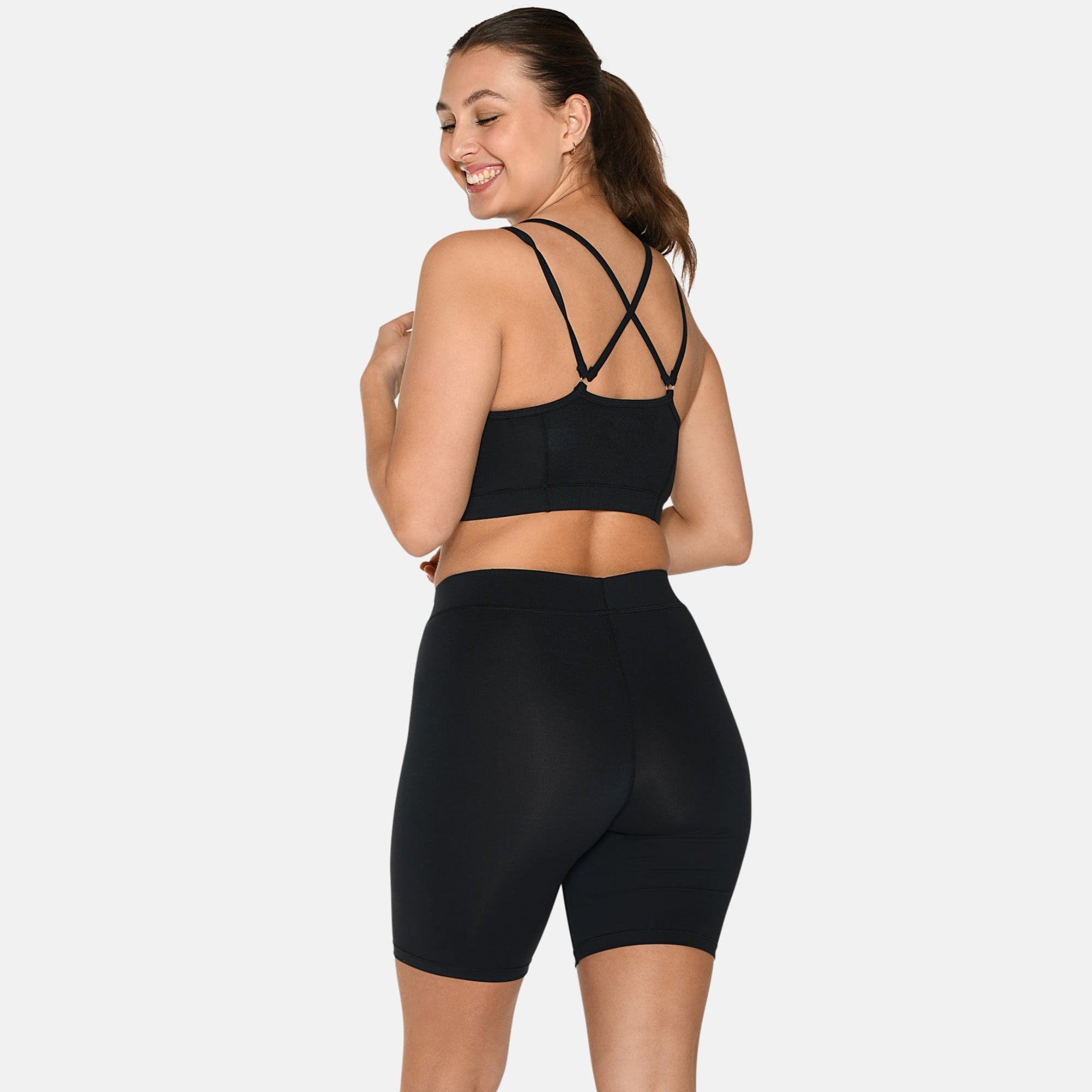 Eine Frau steht lächelnd mit dem Gesicht von der Kamera weg. Sie trägt einen schwarzen Sport-BH mit überkreuzten Trägern und dazu passende hoch taillierte Radlerhosen, gepaart mit weißen Turnschuhen, vor einem schlichten weißen Hintergrund.