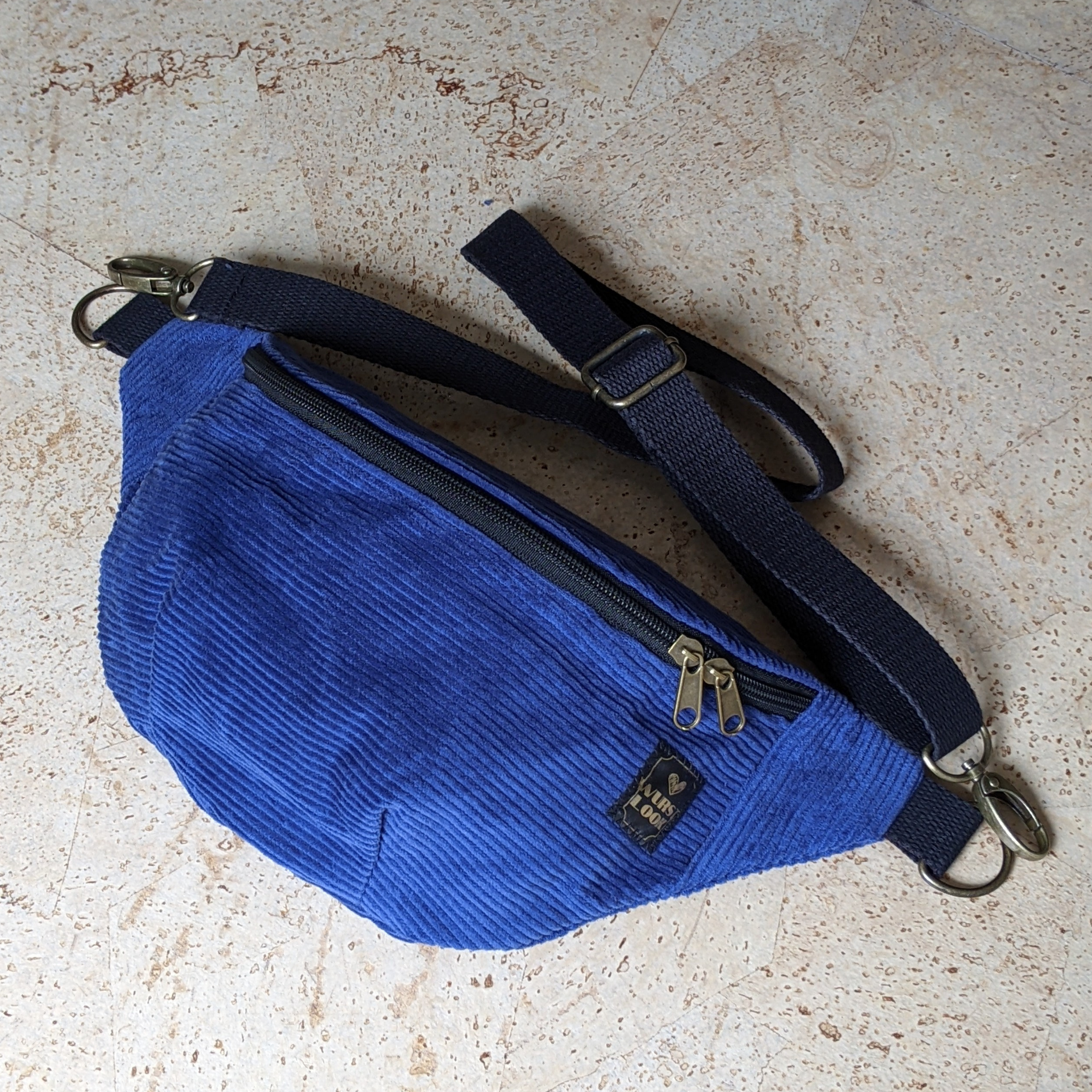 Eine blaue Gürteltasche aus Cord mit einem schwarzen, verstellbaren Riemen, einem goldenen Reißverschluss und einem kleinen, rechteckigen, schwarzen Aufnäher auf der Vorderseite, der auf einer leicht strukturierten Oberfläche angebracht ist.