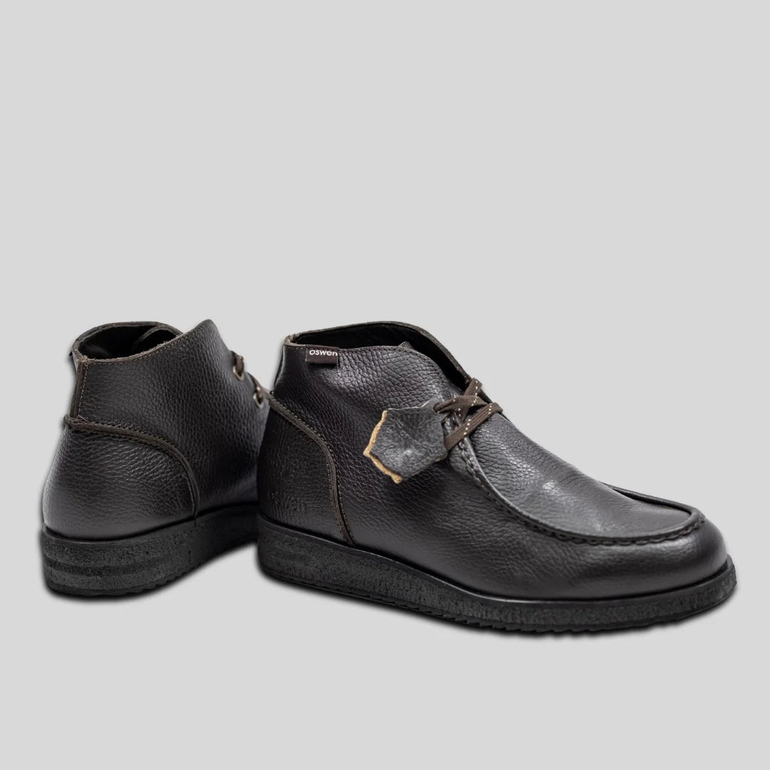 Ein Paar knöchelhohe Schuhe aus dunkelbraunem Leder mit strukturiertem Obermaterial und Gummisohle, wobei ein Schuh nach vorne zeigt und der andere so gedreht ist, dass der Absatz sichtbar ist.