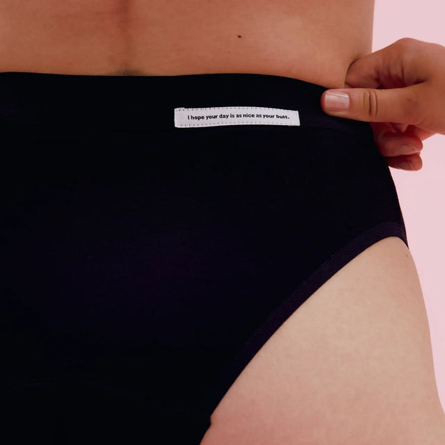 Eine Nahaufnahme einer Person, die schwarze Unterwäsche mit hoher Taille trägt. Auf einem kleinen Etikett am Bund steht: "Ich hoffe, dein Tag ist so schön wie dein Hintern. Der Hintergrund ist hellrosa.