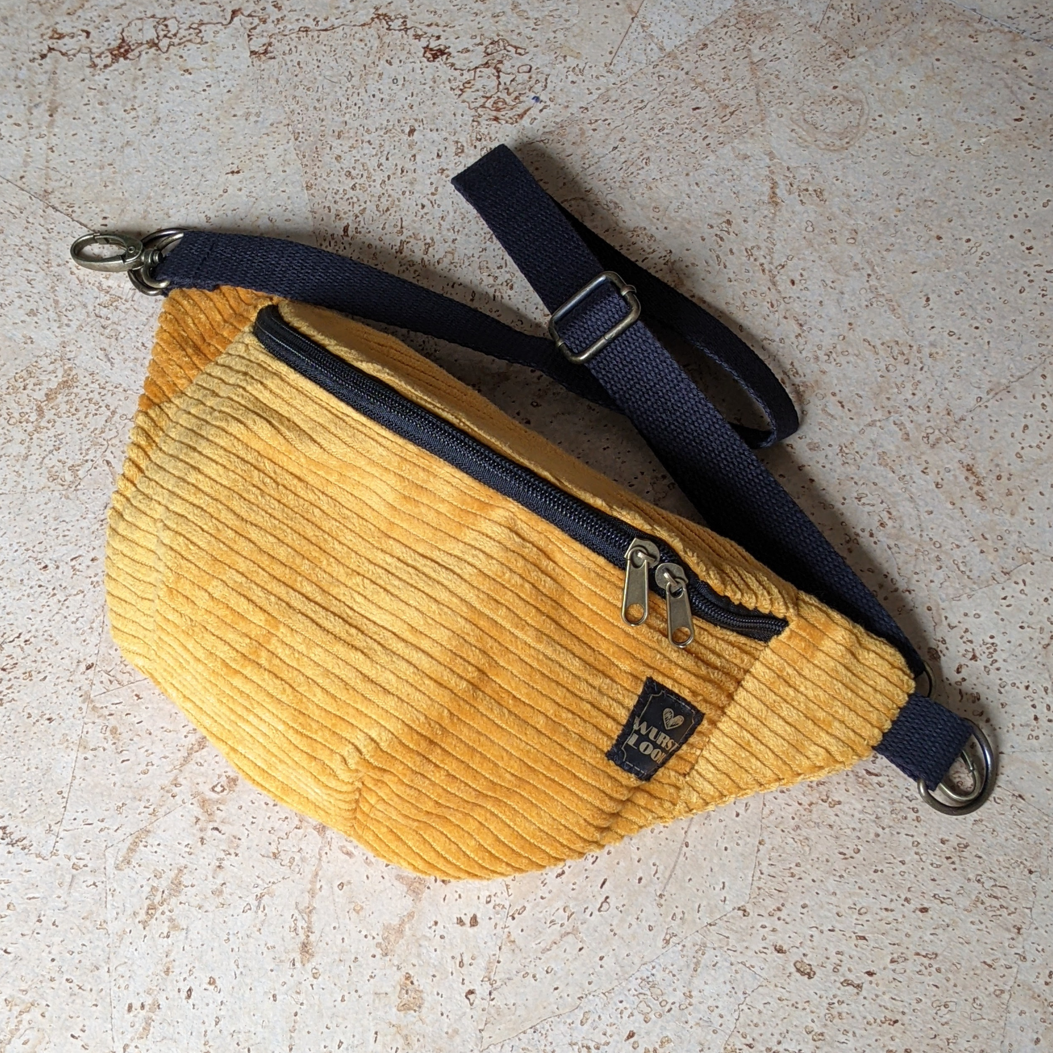 Eine gelbe Gürteltasche aus Cord mit einem schwarzen, verstellbaren Riemen und einer schwarzen Reißverschlusstasche ist auf einer hellen, strukturierten Oberfläche angebracht.