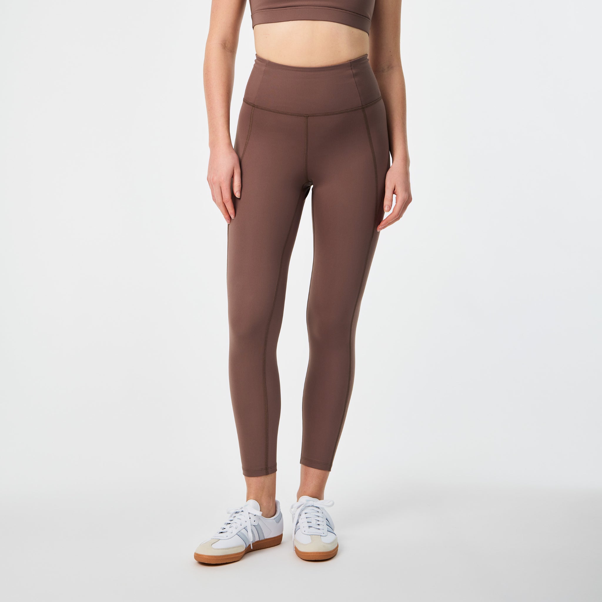 Eine Person, die einen braunen Sport-BH und dazu passende hoch taillierte Leggings trägt, steht vor einem weißen Hintergrund. Ihr Kopf ist nicht zu sehen, und sie trägt weiße Turnschuhe mit hellbraunen Sohlen.