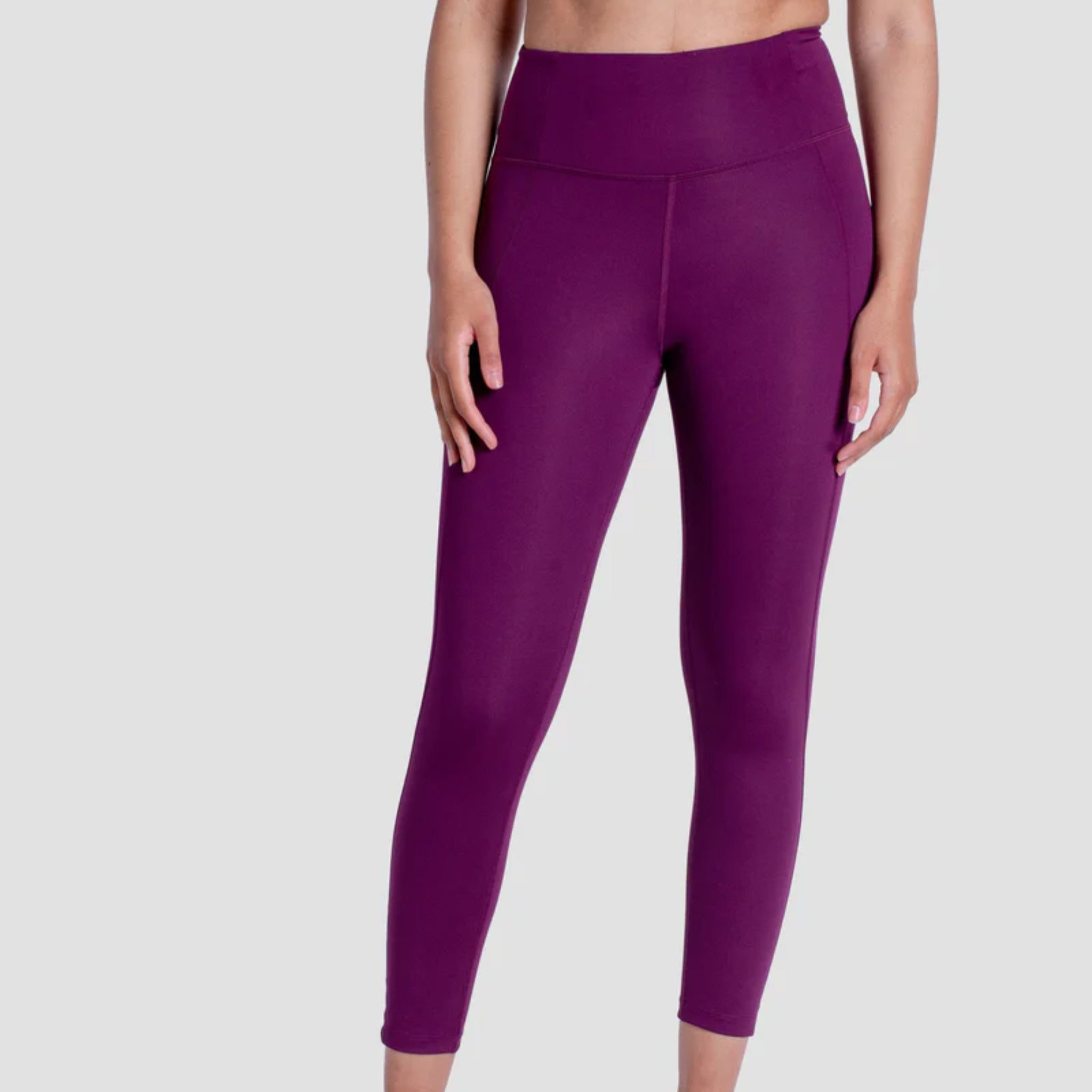Eine Person in knöchellangen, hochgezogenen lila Leggings steht vor einem hellgrauen Hintergrund. Das Bild ist von der Körpermitte bis zu den Füßen abgeschnitten.