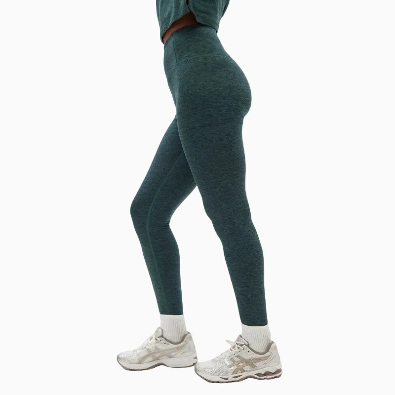 Seitlich stehende Person in hochtaillierten, blaugrünen Leggings, einem abgeschnittenen Oberteil, weißen Socken und weißen Sportschuhen vor einem schlichten weißen Hintergrund. Nur die untere Hälfte des Körpers ist sichtbar.