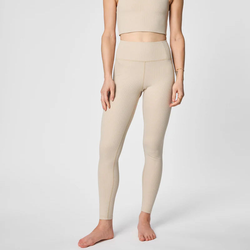 Eine Frau steht barfuß vor einem schlichten Hintergrund und trägt ein passendes beigefarbenes geripptes Crop-Top und ein Set aus hochtaillierten Leggings. Ihre Arme liegen entspannt an den Seiten, und ihr Kopf ist auf dem Bild nicht zu sehen.