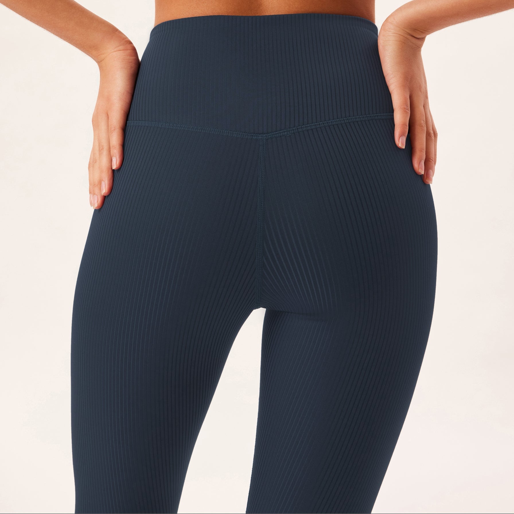 Eine Person, die einen passenden dunkelblauen gerippten Sport-BH und hoch taillierte Leggings trägt, wird von hinten gezeigt, wobei sie die Hände in die Hüften stützt, vor einem hellen Hintergrund.