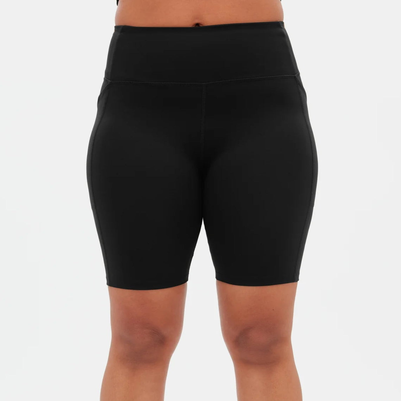 Person, die schwarze, hochtaillierte Radlerhosen, ein schwarzes Crop-Top, weiße Socken und weiße Turnschuhe mit hellbraunen Details trägt und vor einem schlichten weißen Hintergrund steht.