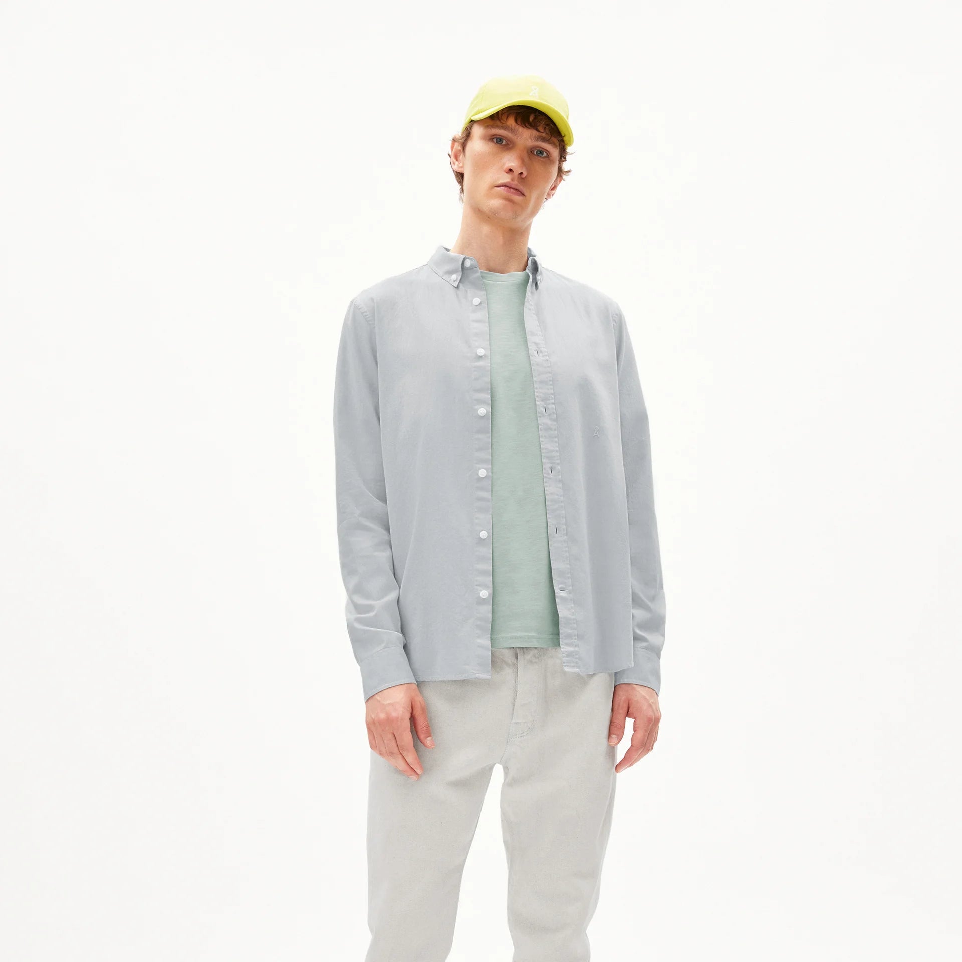 Eine Person steht vor einem weißen Hintergrund und trägt ein hellblaues Button-up-Hemd über einem grünen T-Shirt, eine hellbeige Hose, beige Schuhe und eine gelbe Baseballmütze.