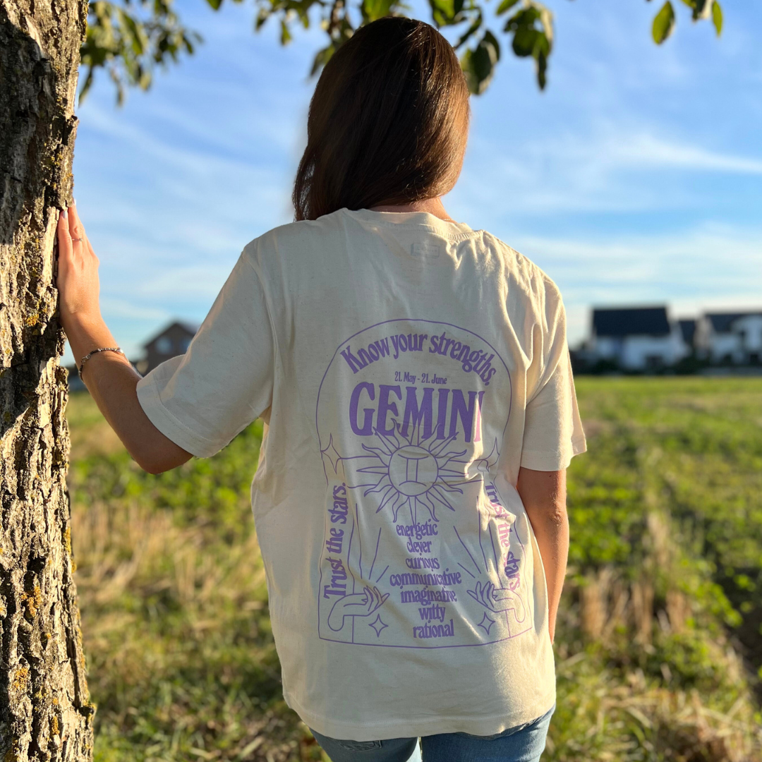 Eine Person steht in einer Landschaft, trägt ein T-Shirt mit "GEMINI" und astrologischen Begriffen.
