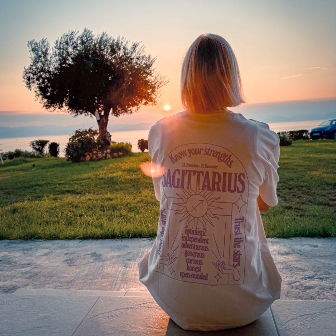 Person sitzt im Sonnenuntergang auf einer Terrasse, trägt ein T-Shirt mit Schütze-Thema und schaut auf das Meer.