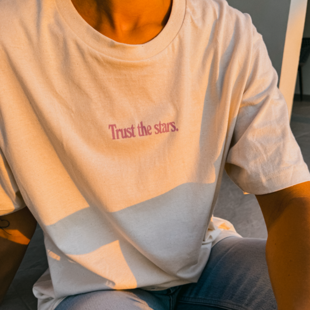 Person trägt ein cremefarbenes T-Shirt mit der Aufschrift "Trust the stars." in lila Schrift.