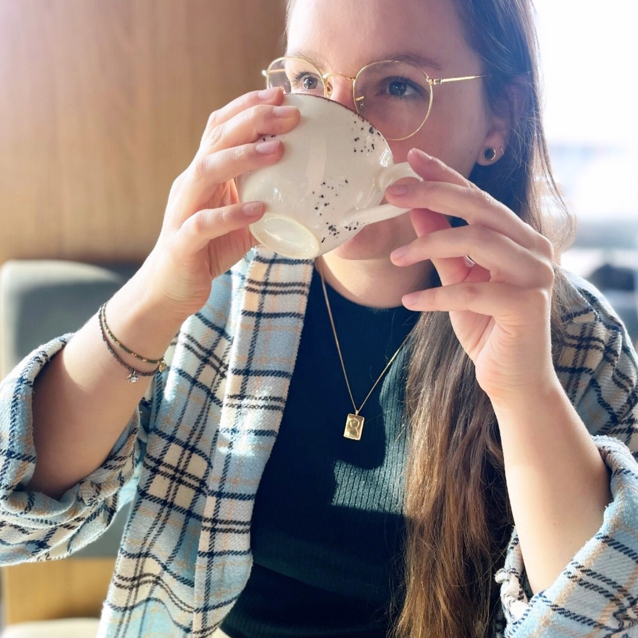 Eine Person mit Brille trinkt aus einer Tasse und trägt eine karierte Jacke und ein schwarzes Oberteil.