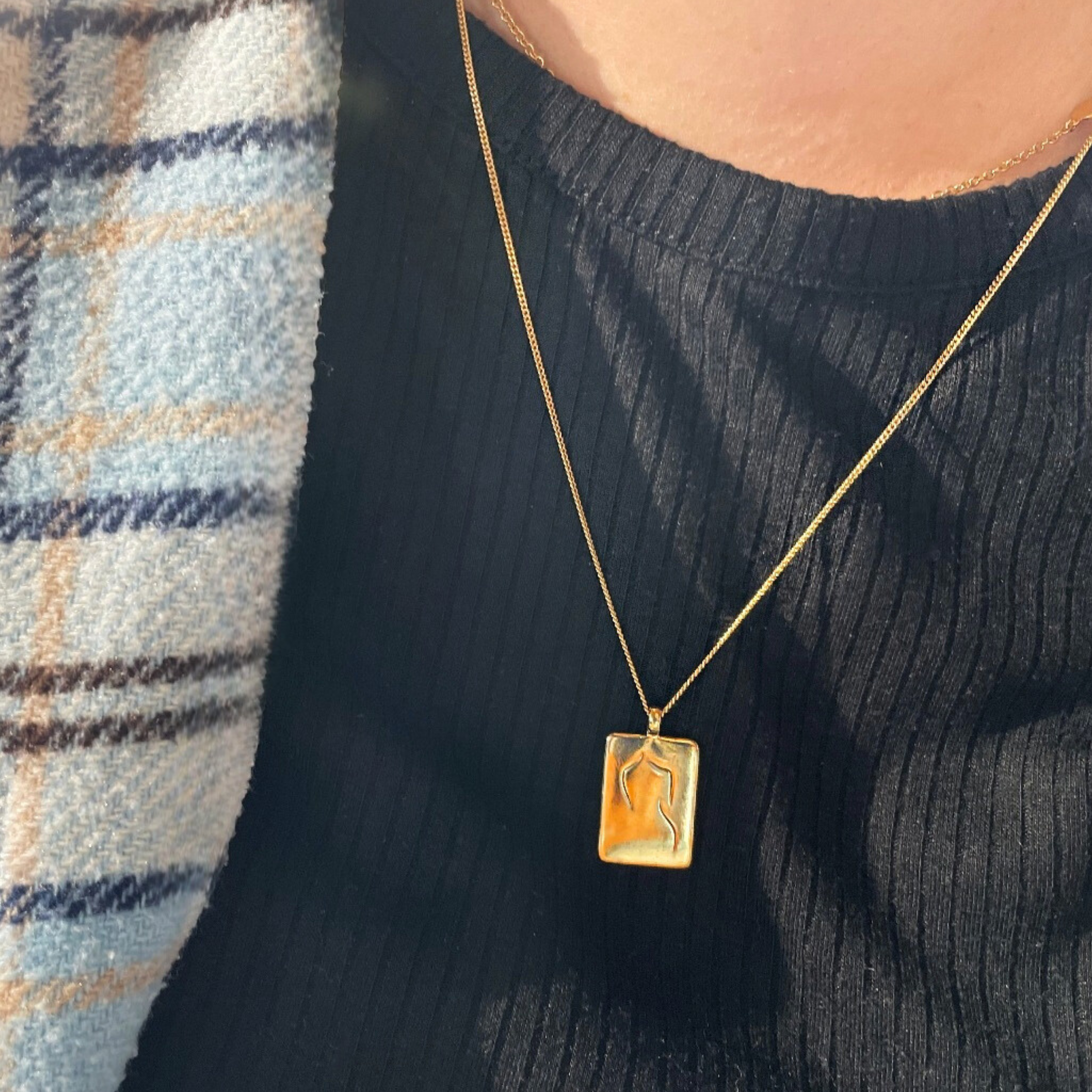Goldene Halskette mit rechteckigem Anhänger auf schwarzem Oberteil neben einem blau karierten Schal.