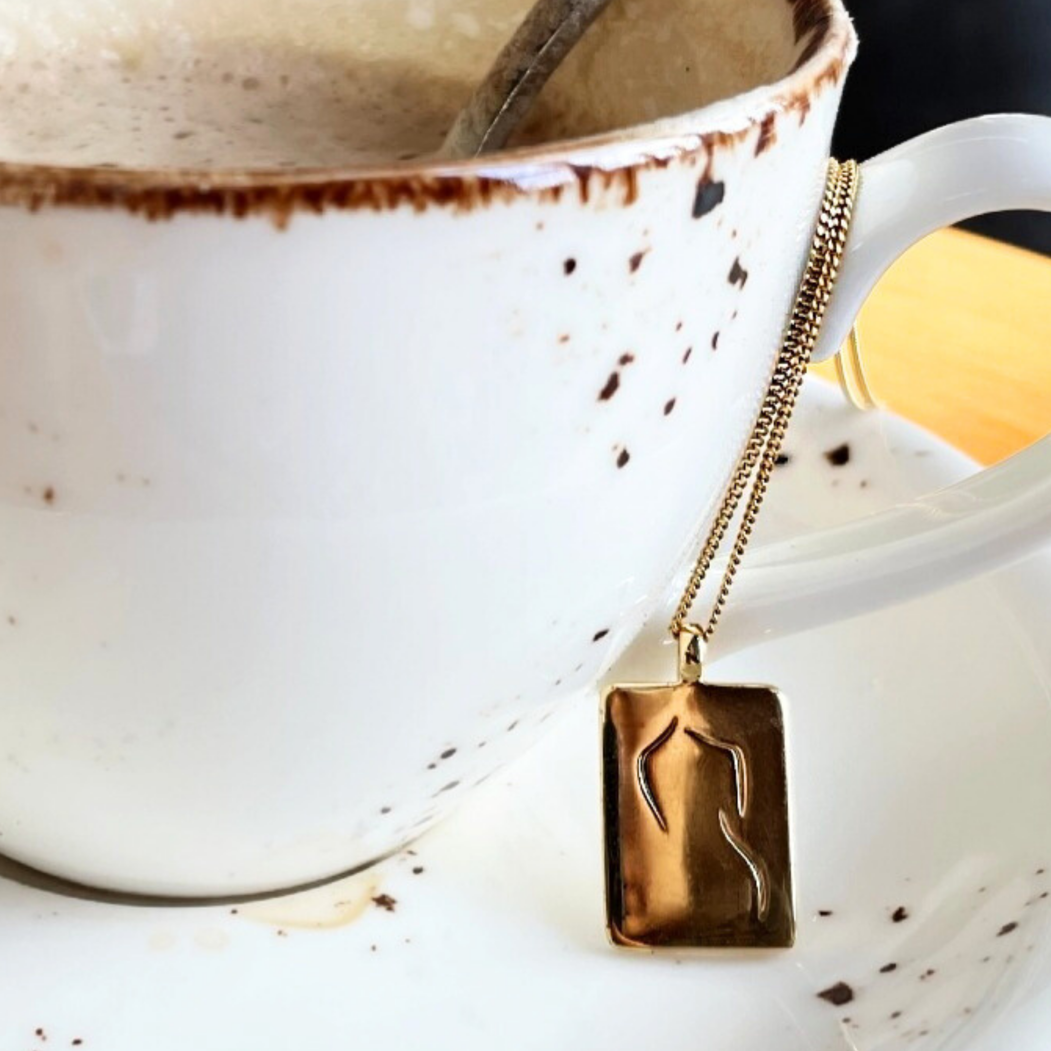 Eine Tasse Kaffee mit Löffel und eine goldene Halskette mit rechteckigem Anhänger hängen am Griff.