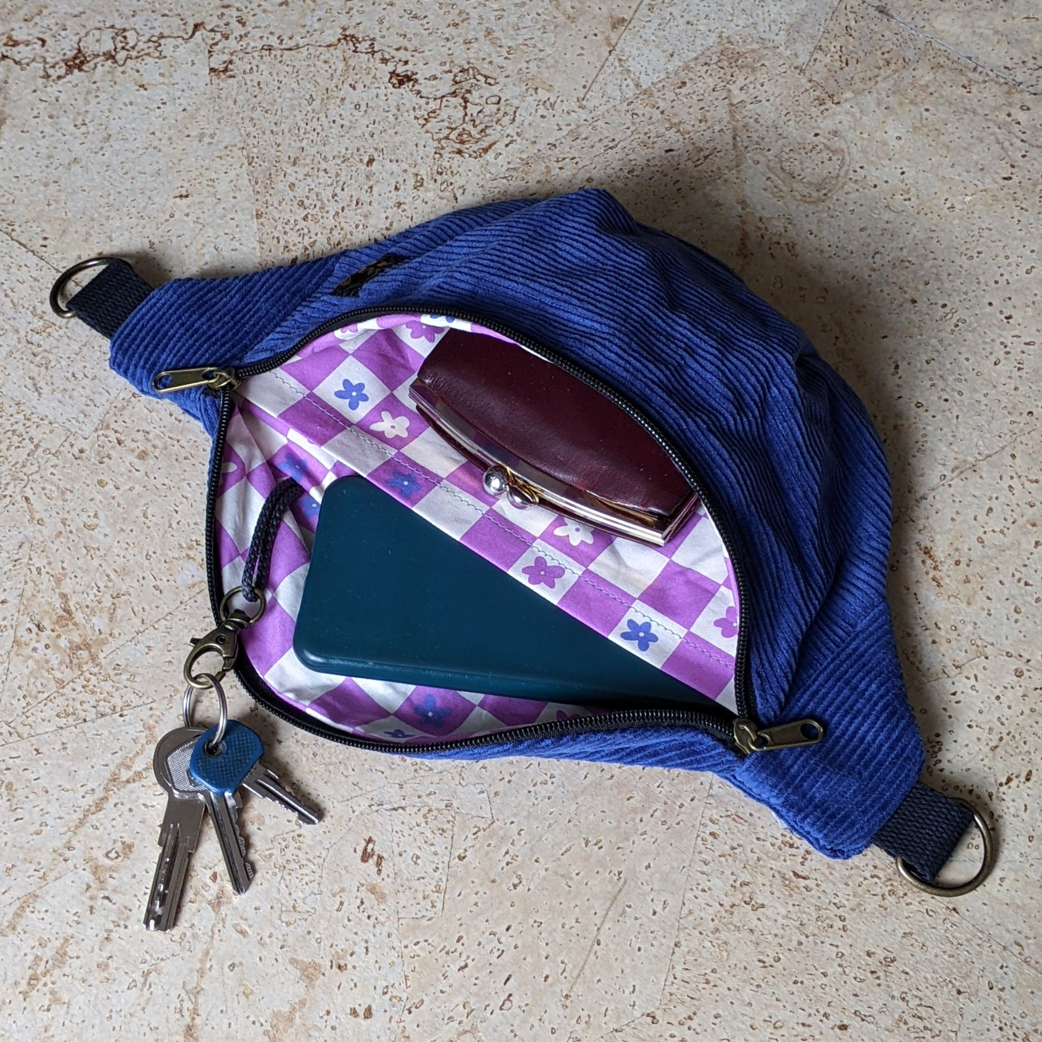 Eine blaue Cordtasche mit Reißverschluss ist geöffnet und offenbart ein kastanienbraunes Portemonnaie, eine blaugrüne Brieftasche und einen Schlüsselbund. Das Futter der Tasche ist ein rosa und weiß kariertes Blumenmuster. Die Tasche ruht auf einer beigen Oberfläche.