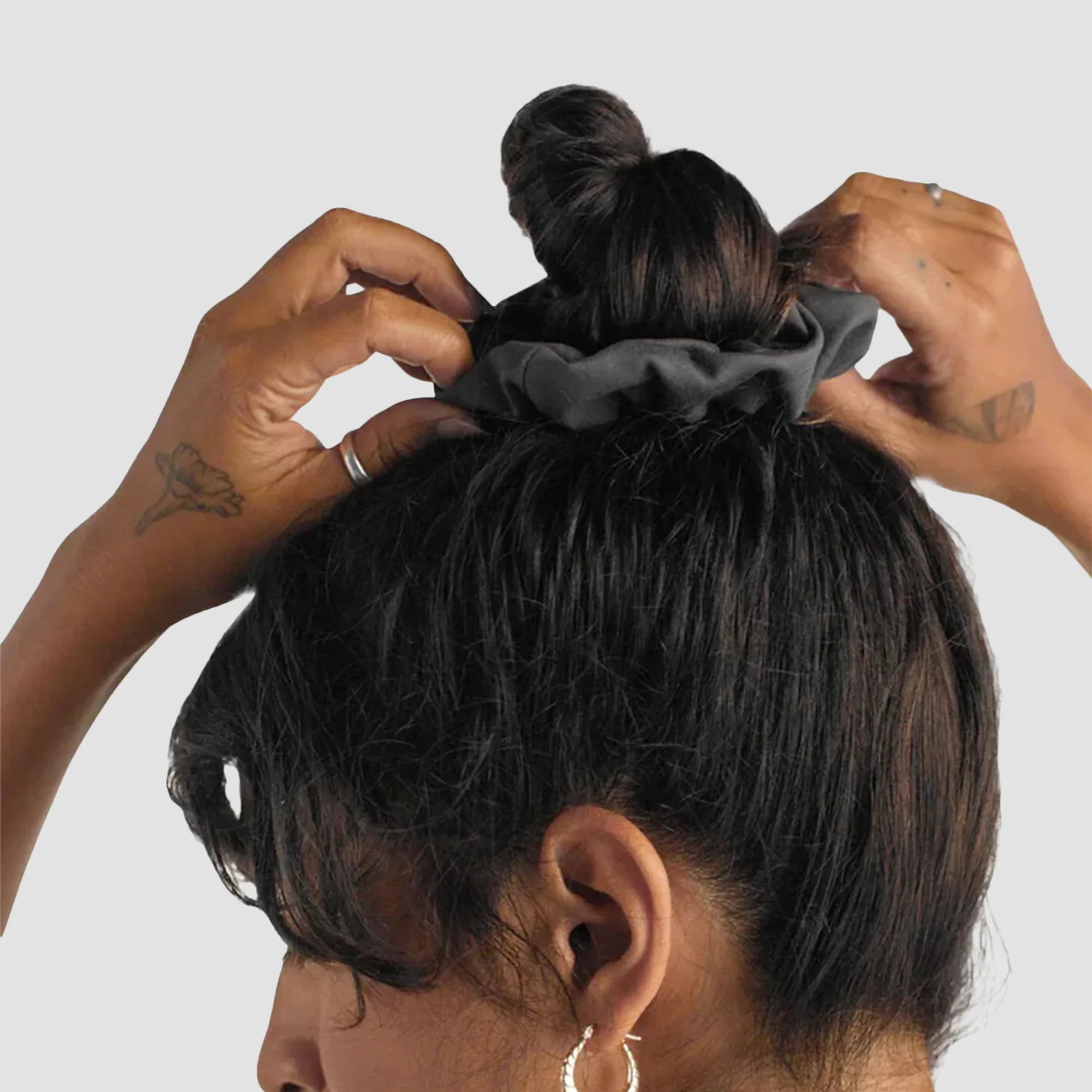 Eine Person mit dunklen Haaren steckt sich ein schwarzes Haargummi in den Dutt. Sie trägt Ohrringe und ein Tanktop, auf ihren Händen sind Tätowierungen zu sehen. Der Hintergrund ist einfarbig hellgrau.