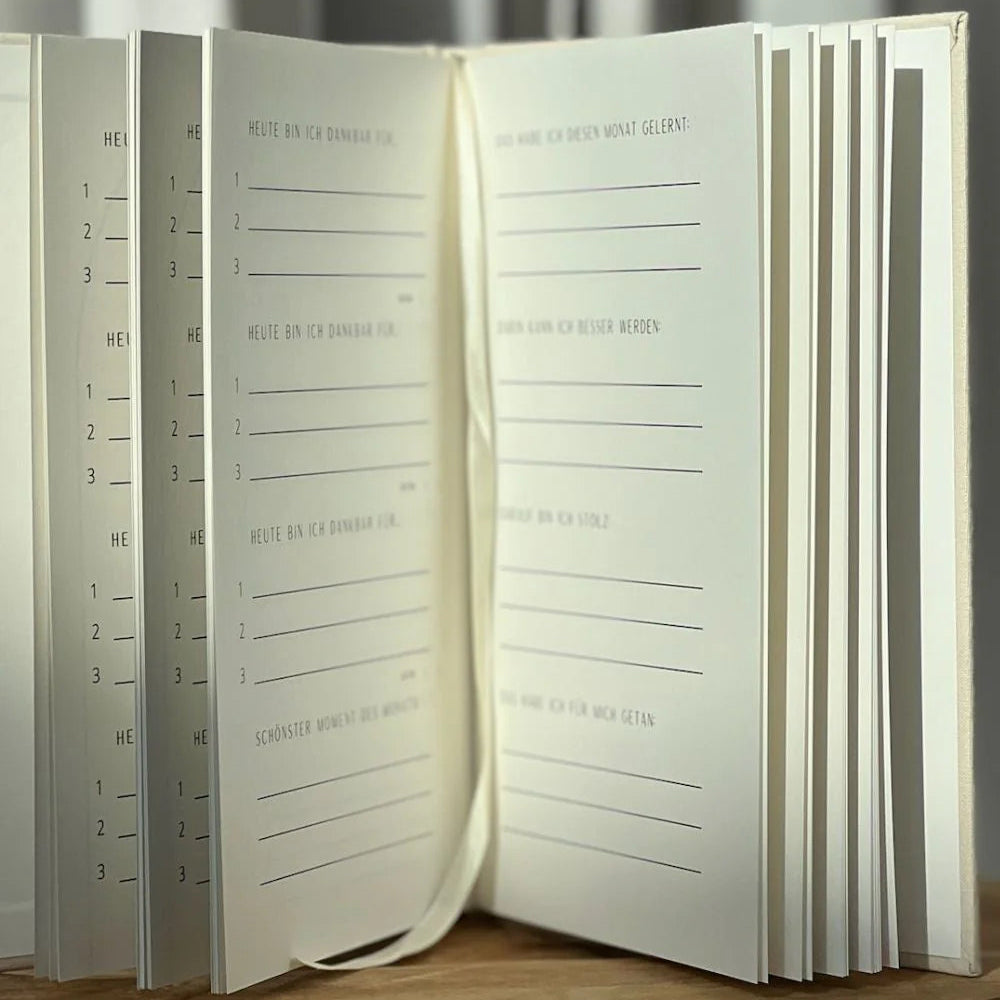 Ein aufgeschlagenes Tagebuch mit linierten Seiten liegt auf einer Holzunterlage. Die Seiten sind in deutscher Sprache mit Aufforderungen und nummerierten Abschnitten für die tägliche Reflexion bedruckt. Sanftes Licht und neutrale Vorhänge schaffen einen ruhigen Hintergrund.
