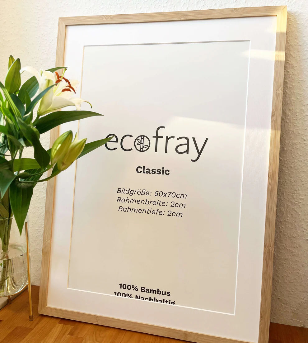 Ein holzgerahmtes Plakat mit der Aufschrift "ecofray Classic" und Angaben in deutscher Sprache steht auf einer Holzfläche neben einer Vase mit weißen Lilien und Grünpflanzen an einer hellen Wand.