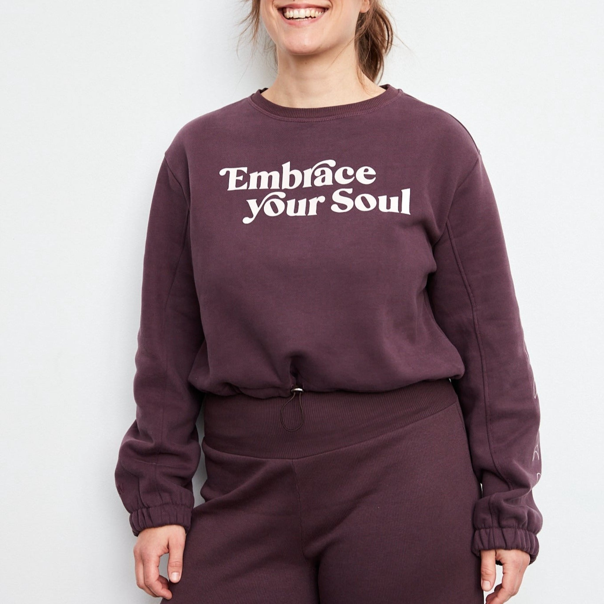 Frau trägt einen lila Pullover mit der Aufschrift "Embrace Your Soul" und passende lila Hose, sie lächelt.