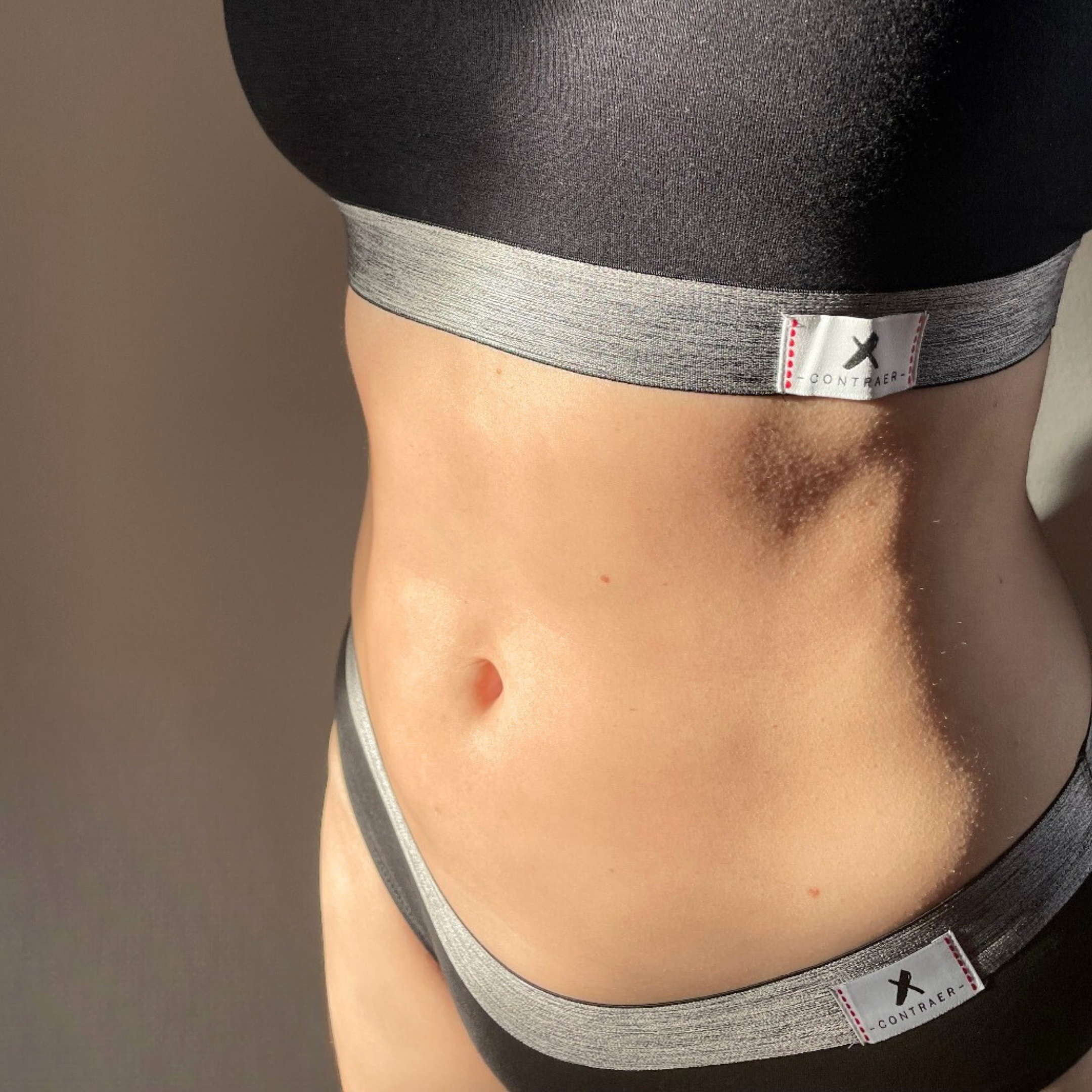 Ausschnitt eines weiblichen Torsos in grauer Sportbekleidung mit Etikett im Sonnenlicht.