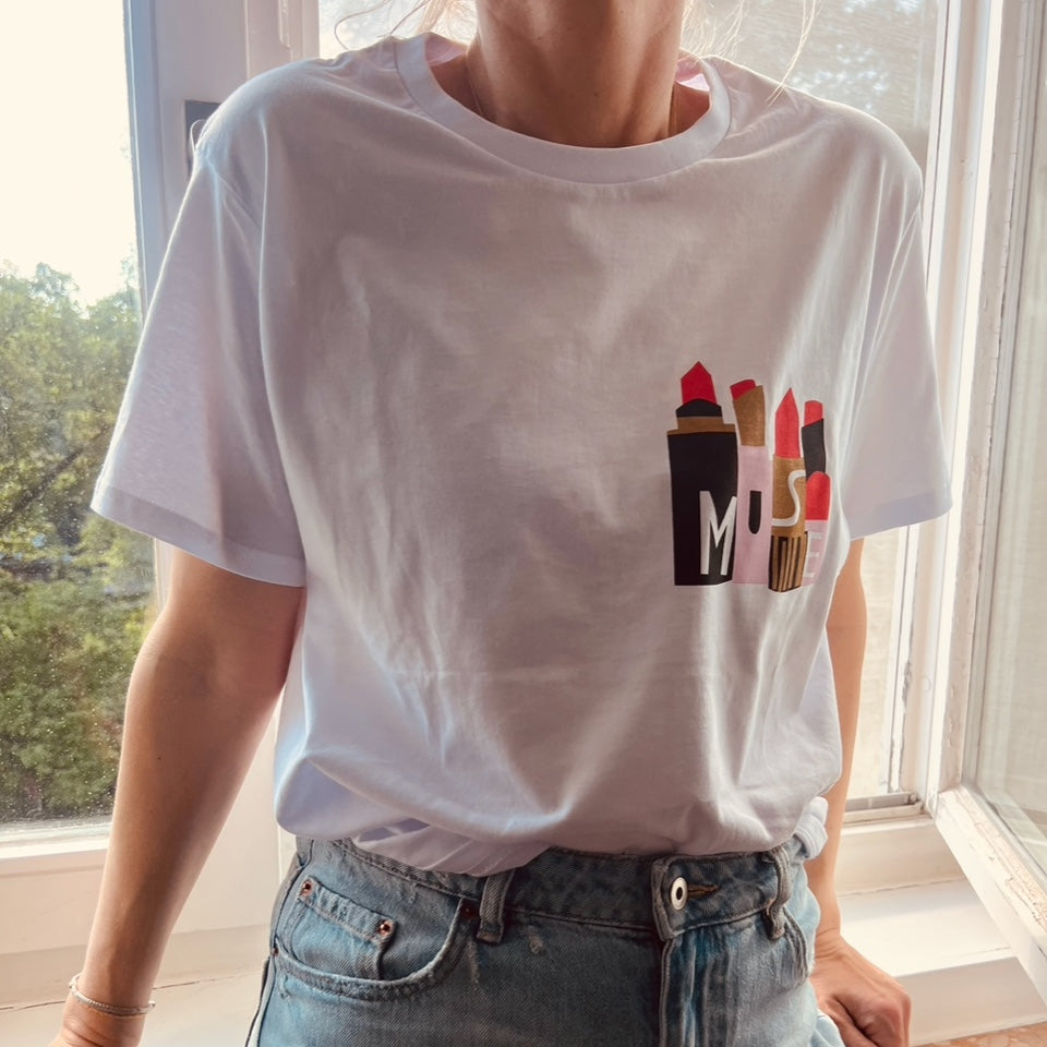 Eine Person, die ein weißes T-Shirt mit bunten Lippenstiftillustrationen auf der Brust trägt, steht vor einem Fenster mit Grünpflanzen draußen. Die Person trägt außerdem eine hellblaue Jeans.
