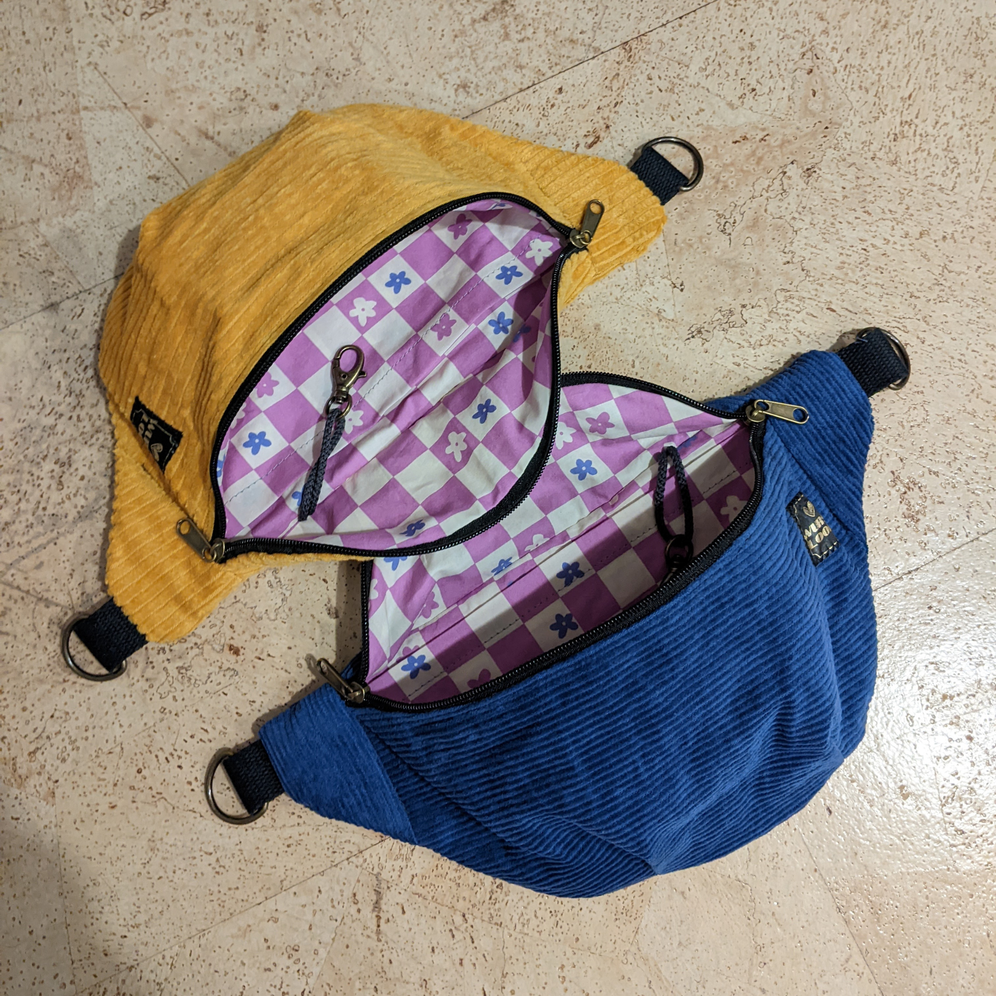 Zwei Gürteltaschen aus Kord, eine gelbe und eine blaue, stehen auf einem beigen Boden. Beide sind geöffnet und offenbaren ein rosa-weiß kariertes Futter mit kleinen lila und blauen Blumen sowie einen Schlüsselclip aus Metall in jeder Tasche.