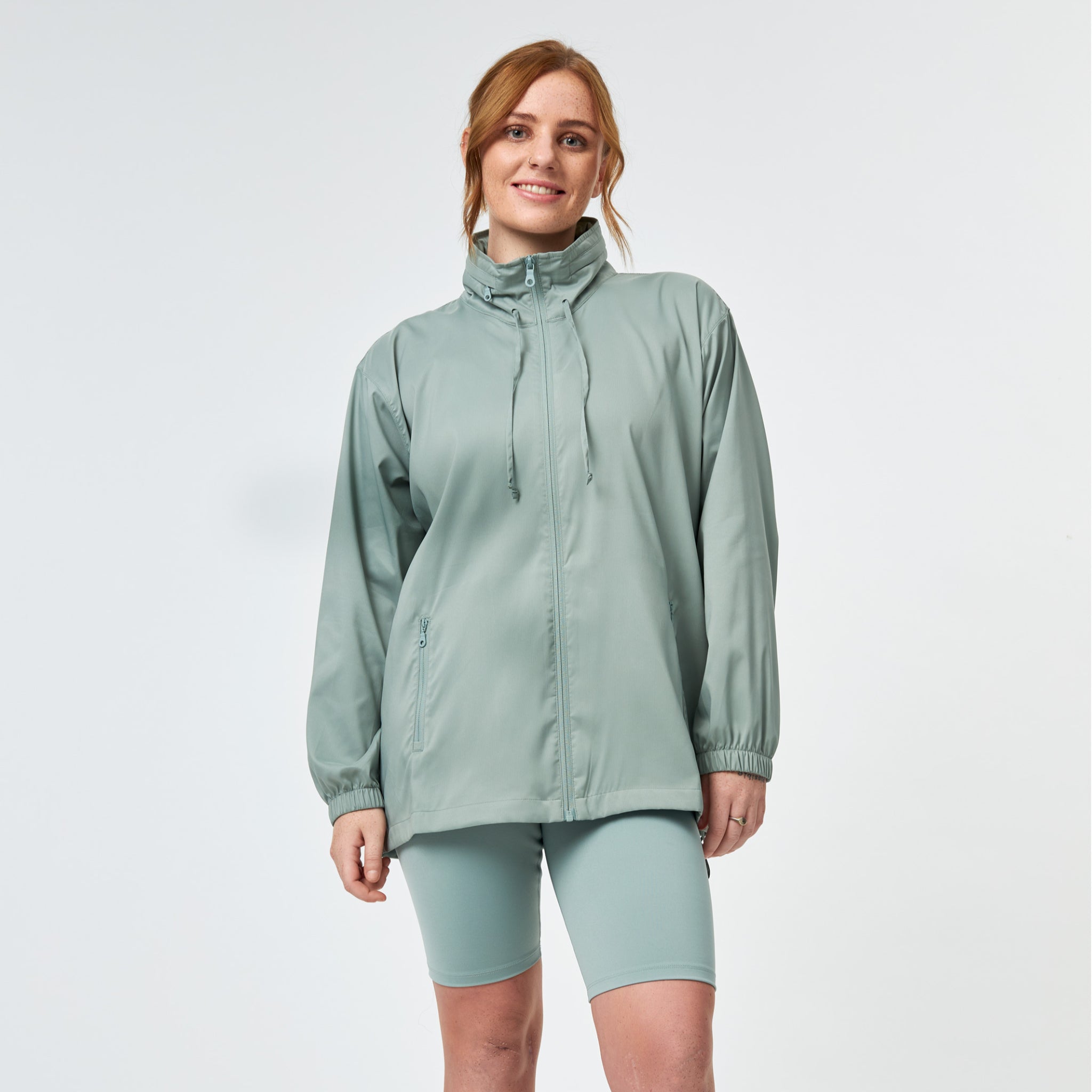 Eine Frau mit roten Haaren, die lächelt und eine hellgrüne Jacke mit Reißverschluss und passende Biker-Shorts trägt, steht vor einem schlichten weißen Hintergrund.