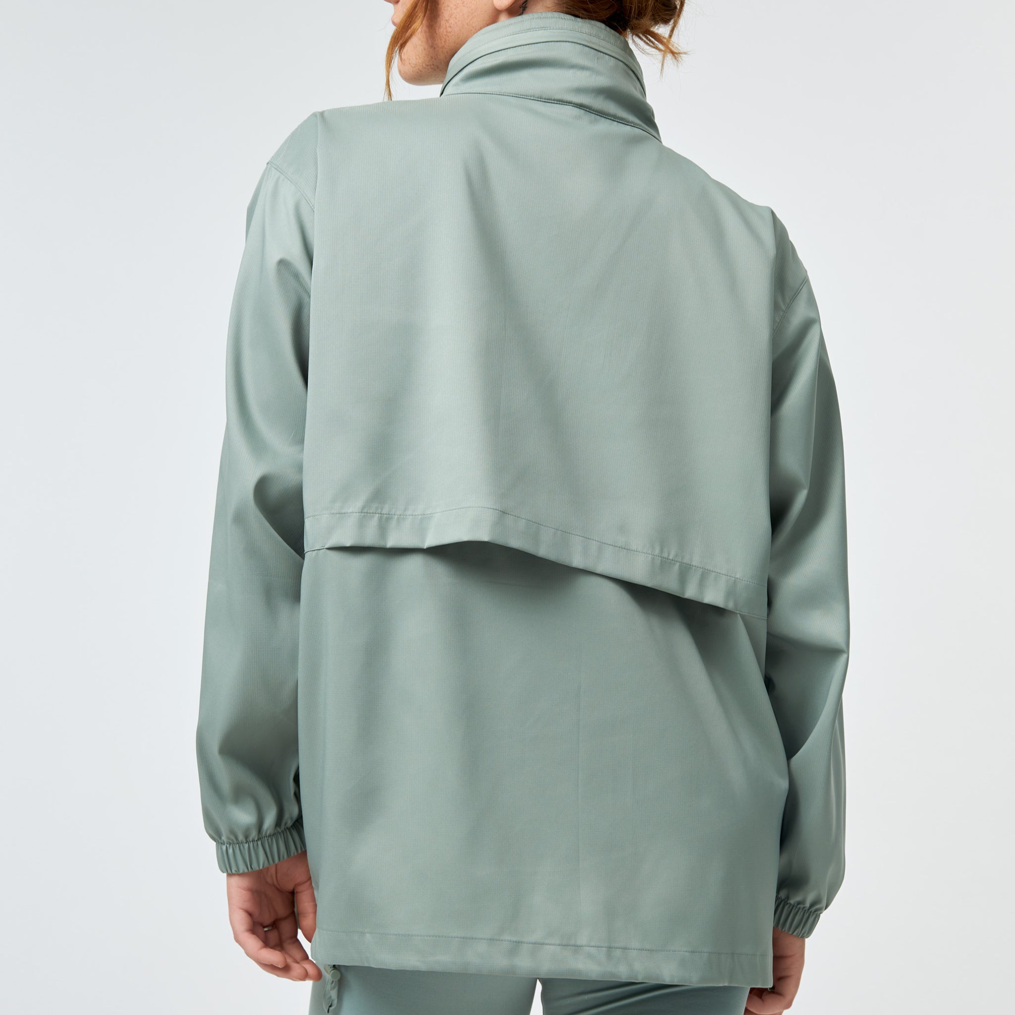 Eine Person mit heller Haut und rötlichem Haar trägt eine lockere, hellsalbeigrüne Jacke und eine dazu passende Hose, die von hinten vor einem schlichten hellen Hintergrund gezeigt wird.