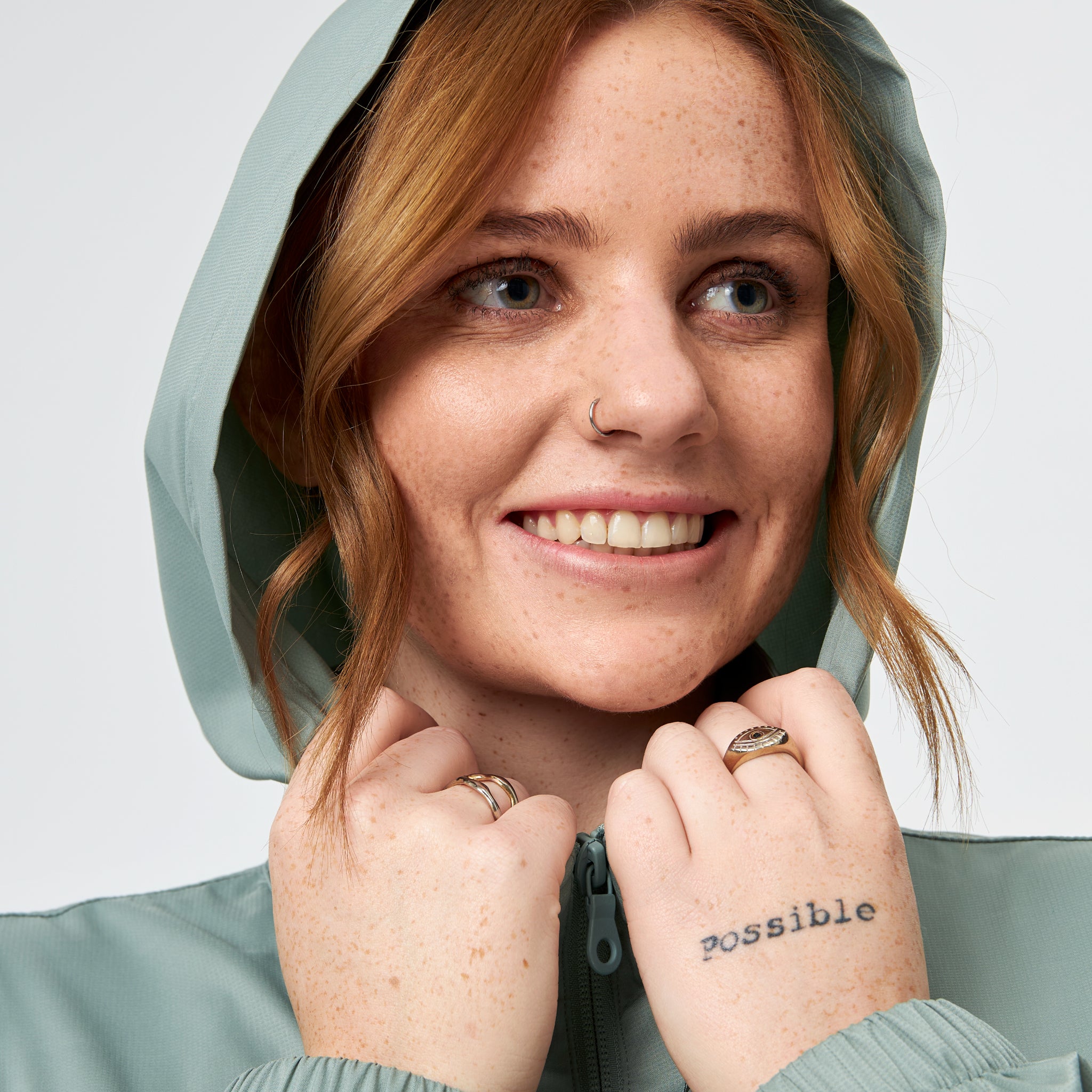 Eine lächelnde Person mit heller Haut, Sommersprossen und kastanienbraunem Haar trägt eine grüne Kapuzenjacke. Sie hält die Kapuze mit beiden Händen und zeigt Ringe und eine mögliche Tätowierung auf ihrer Hand.