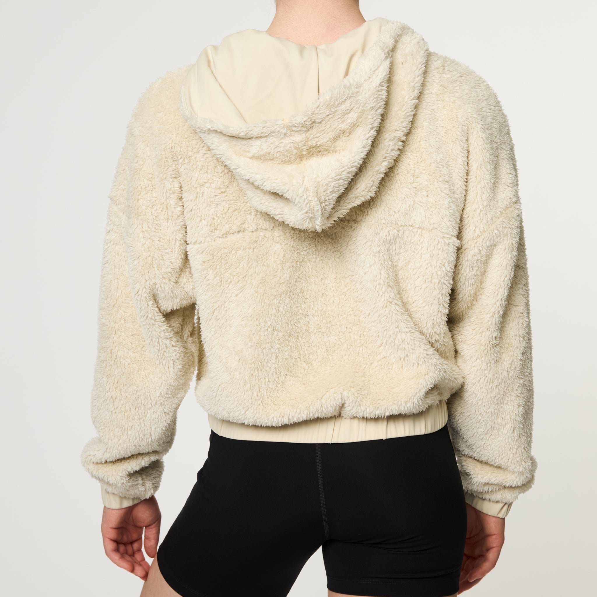 Eine Person, die eine cremefarbene, wattierte Kapuzenjacke und schwarze Shorts trägt, wird von hinten gezeigt und steht vor einem schlichten hellen Hintergrund.