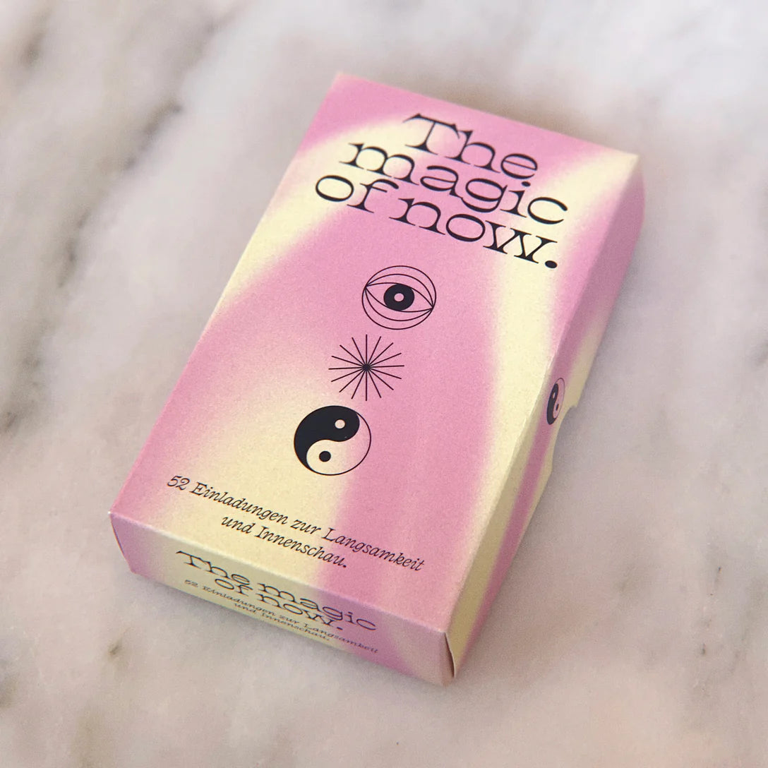 Eine Schachtel mit rosa und gelbem Farbverlauf mit dem Titel Die Magie des Jetzt. mit mystischen Symbolen, darunter ein Auge, ein Sternenhimmel und ein Yin-Yang. Die Box enthält 52 Karten für Achtsamkeit und Introspektion in deutscher Sprache.