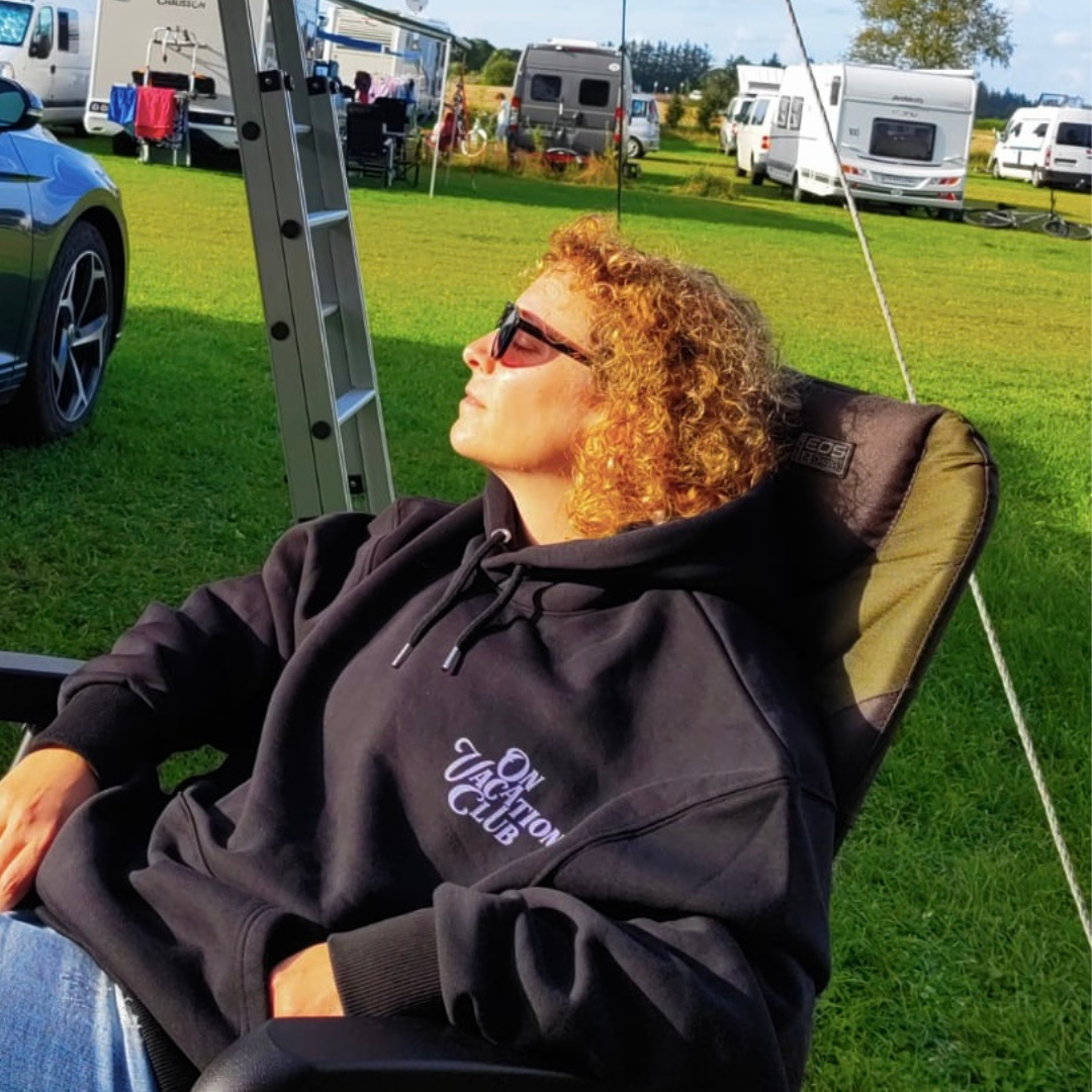 Frau mit Sonnenbrille und Kapuzenpullover entspannt auf einem Campingstuhl, umgeben von Wohnwagen.