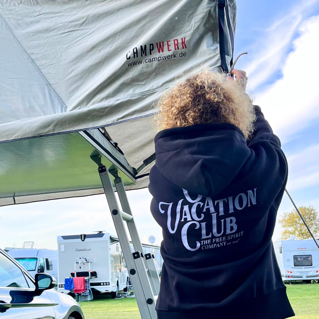 Person dörrt ein Zelt an einem Campingplatz auf, mit Wohnmobilen im Hintergrund.
