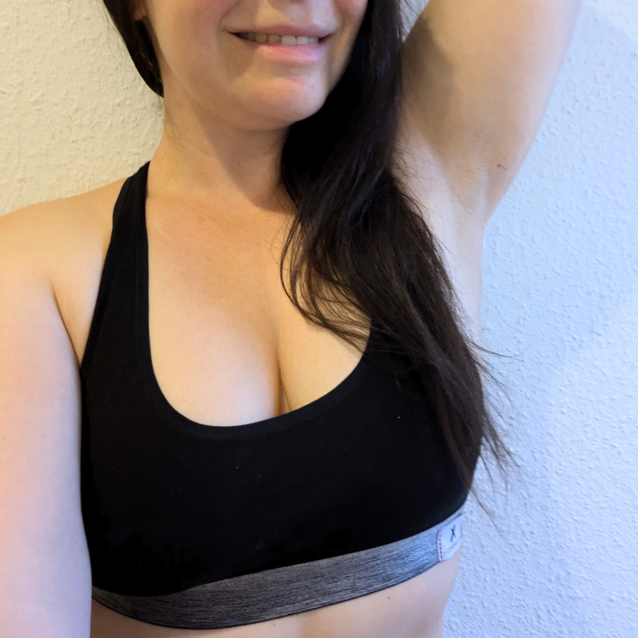 Eine Frau in einem schwarzen Sport-BH lächelt und hebt einen Arm, wodurch ein Teil ihres Gesichts und lange Haare sichtbar sind.