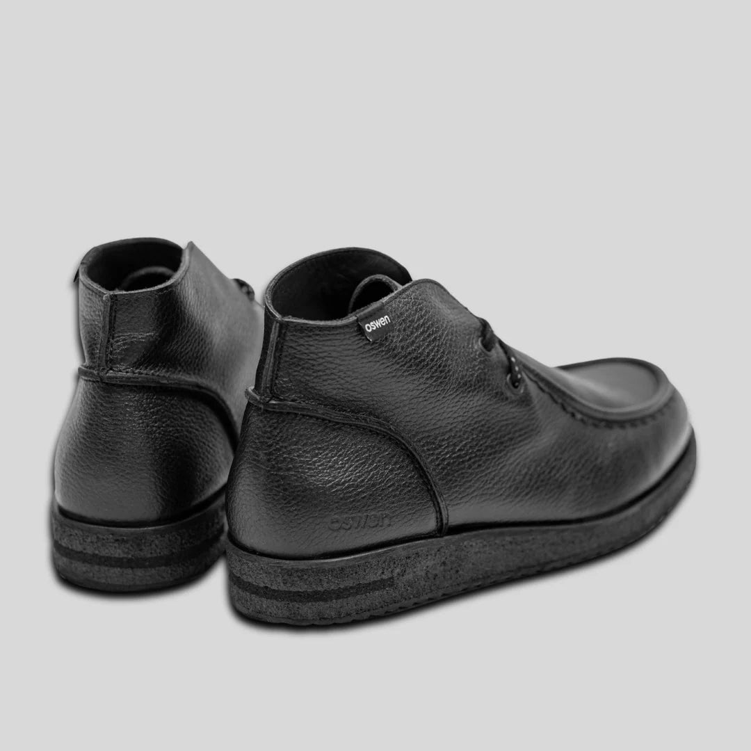 Ein Paar schwarzer Lederknöchelschuhe mit strukturierten Sohlen, dargestellt vor einem schlichten hellgrauen Hintergrund. Die Schuhe sind so positioniert, dass die Absätze leicht nach außen zeigen.