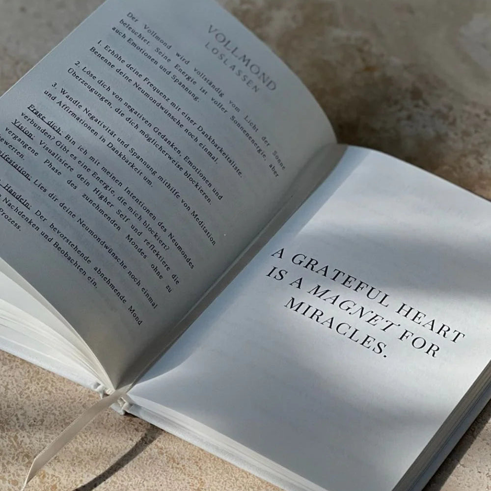 Ein aufgeschlagenes Buch liegt auf einer hellen Fläche. Auf der einen Seite steht ein Text in deutscher Sprache, auf der anderen: EIN GUTES HERZ IST EIN MAGNET FÜR WUNDER. Das Sonnenlicht wirft weiche Schatten auf das Buch.
