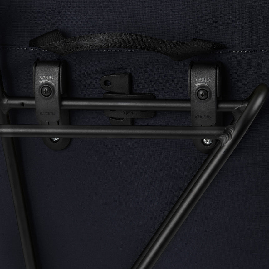 Nahaufnahme eines schwarzen Metallträgers mit zwei Kunststoffklammern mit der Aufschrift Vario, der an einer dunklen Stoffoberfläche befestigt ist und wahrscheinlich zu einem Befestigungssystem für Fahrradtaschen gehört.